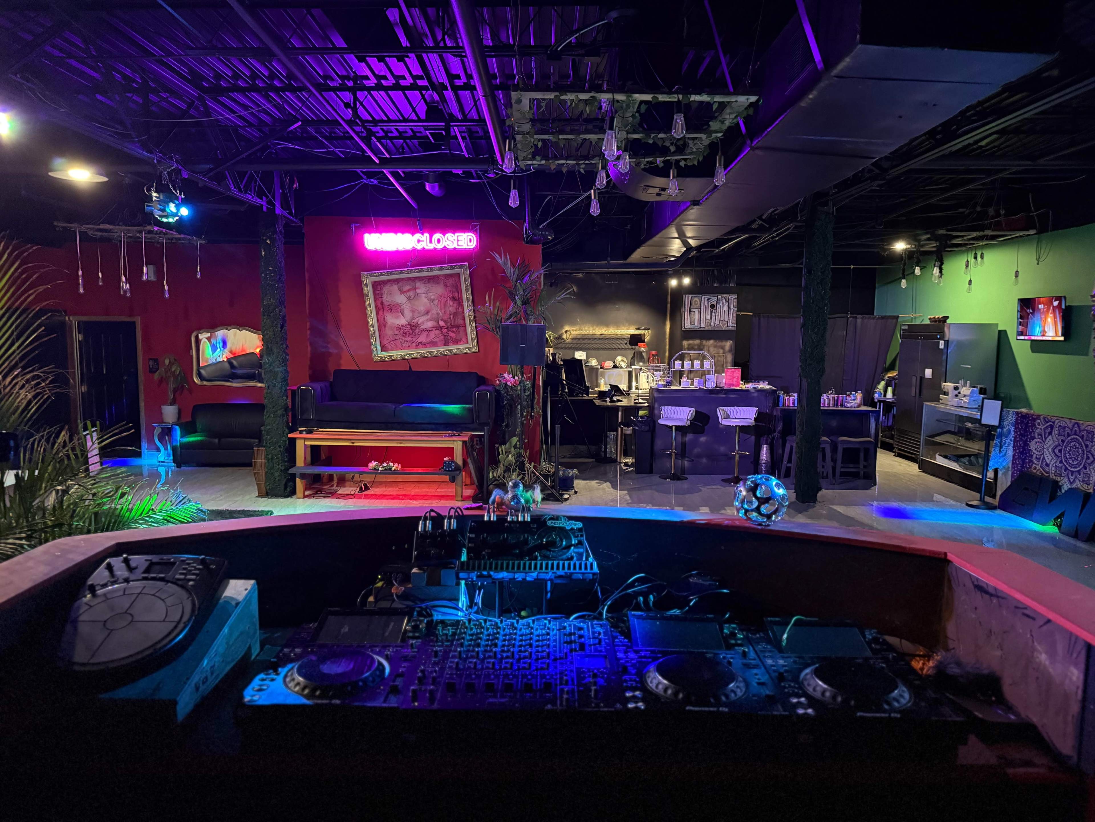 The image shows an indoor venue with vibrant lighting, a DJ booth in the foreground, and lounge areas in the background featuring a bar and small tables.