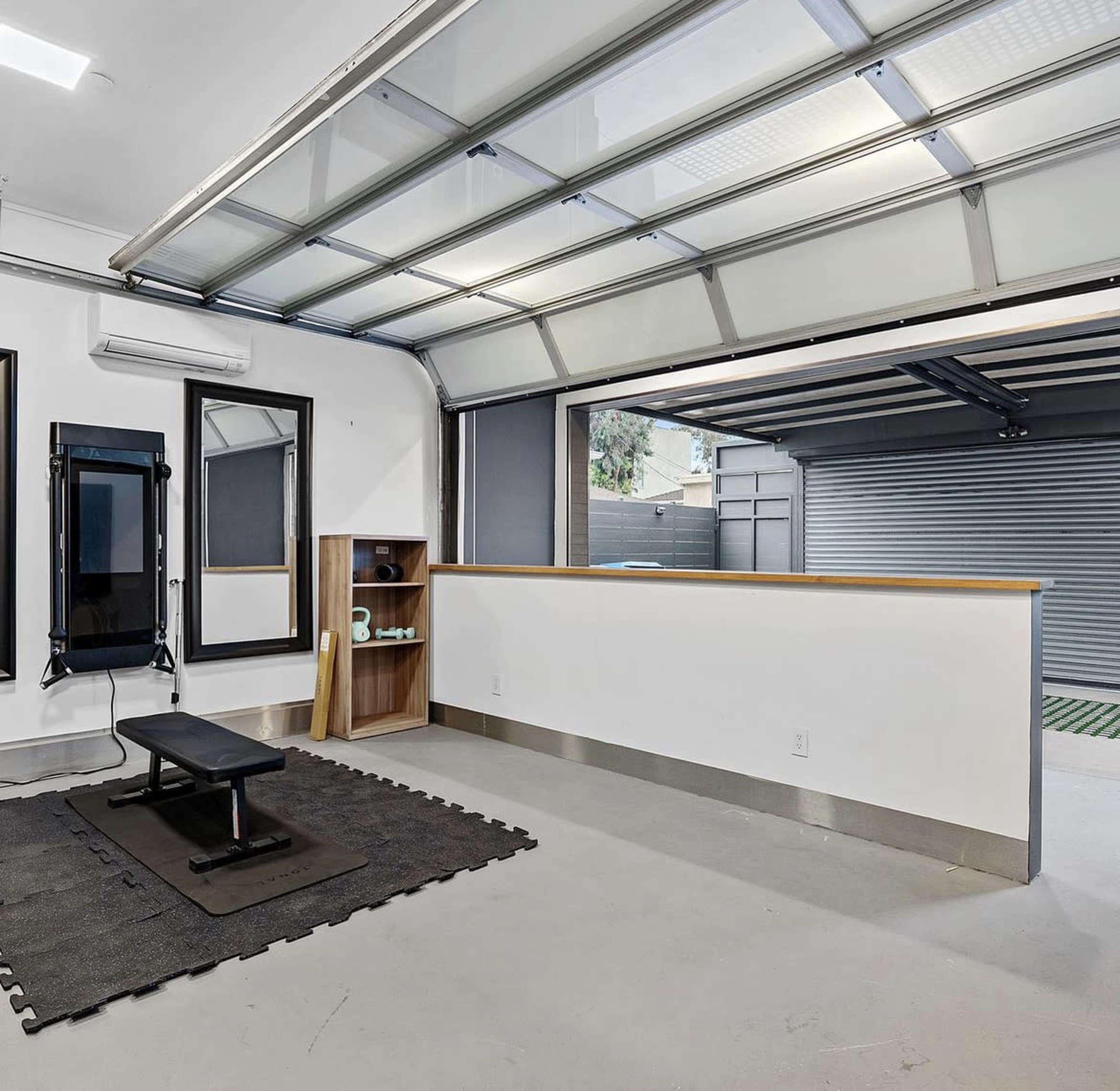 A modern garage with an open door revealing a view of an outdoor area, featuring a gym setup and a storage shelf against a white wall.