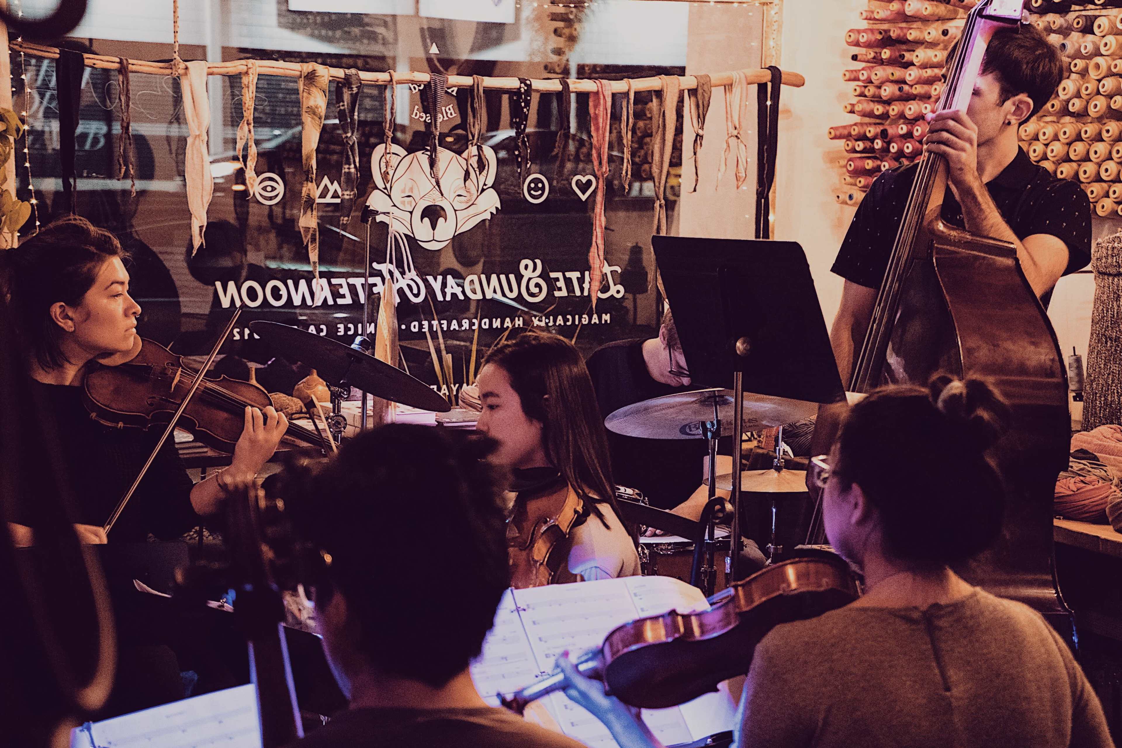 A group of musicians plays various instruments in a cozy setting, with stringed instruments and a drum kit visible.