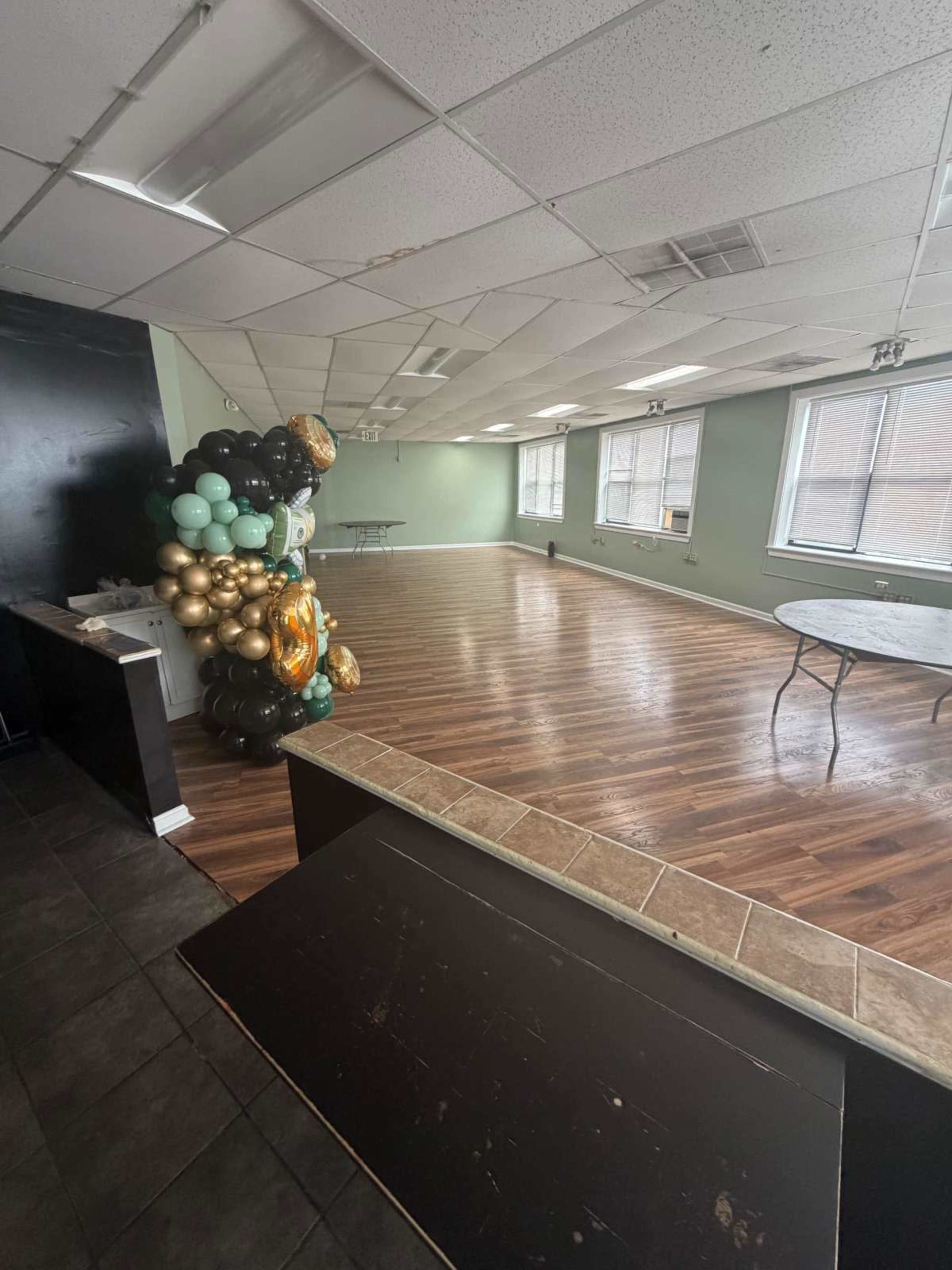 A large, empty event space features wooden flooring, green walls, and several tables set up along the sides, with a decorative balloon arrangement in the corner.