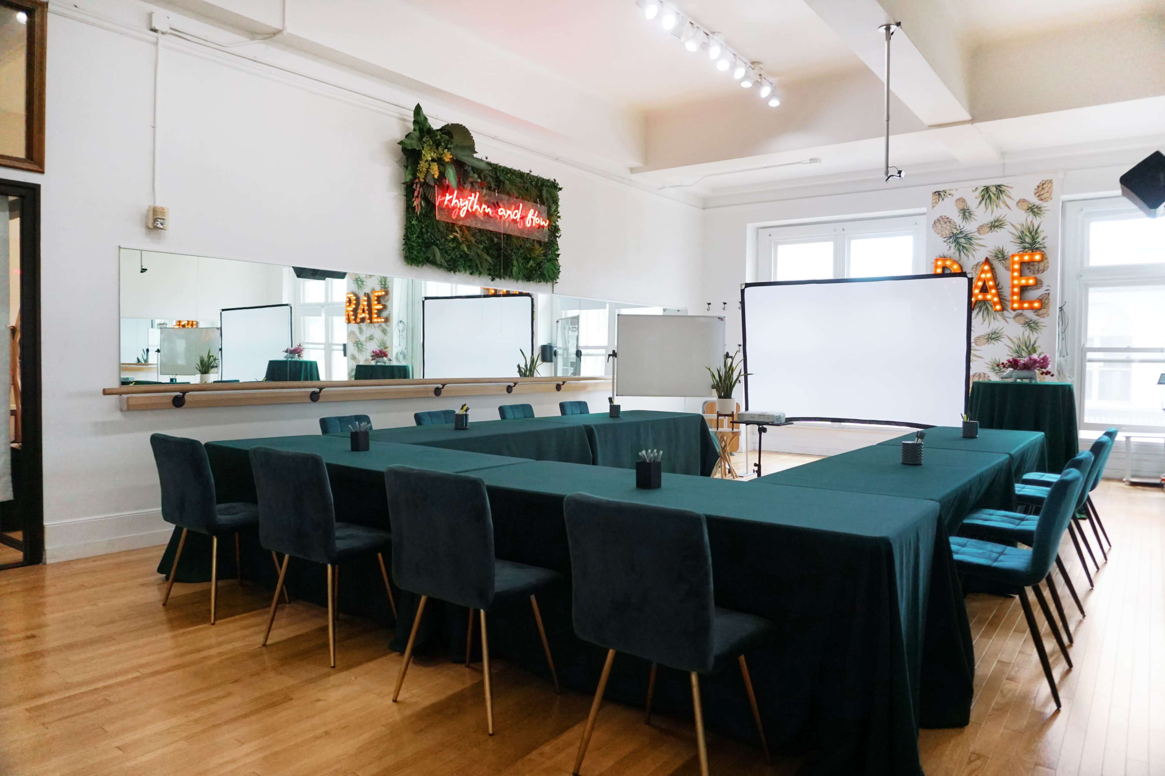 A conference room is set up with a long table covered in green cloth and surrounded by chairs, featuring a neon sign and decorative plants on the walls.