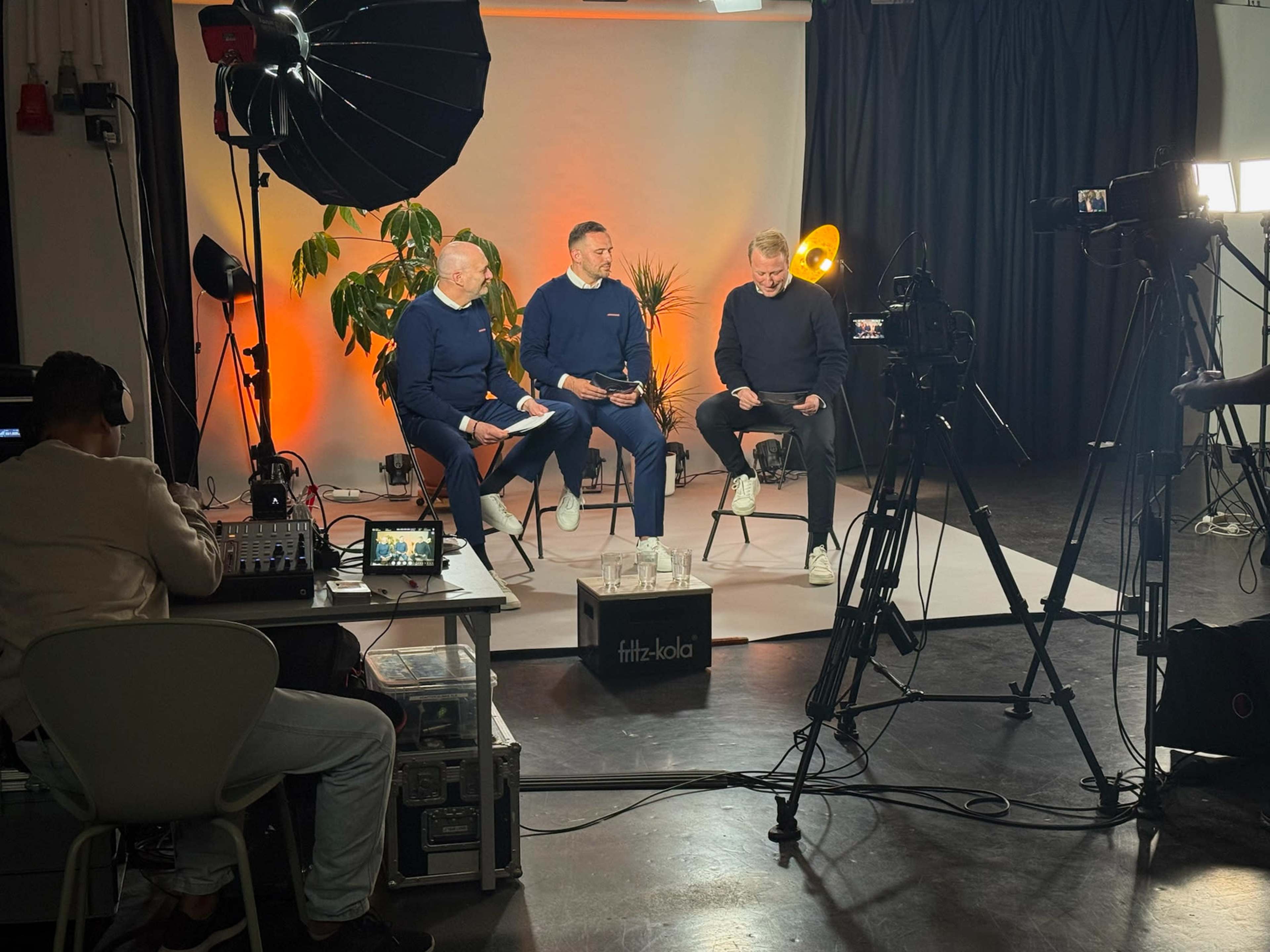 Three men in matching outfits sit on stools, discussing in a studio setup with cameras and lighting equipment, while a person operates a soundboard nearby.