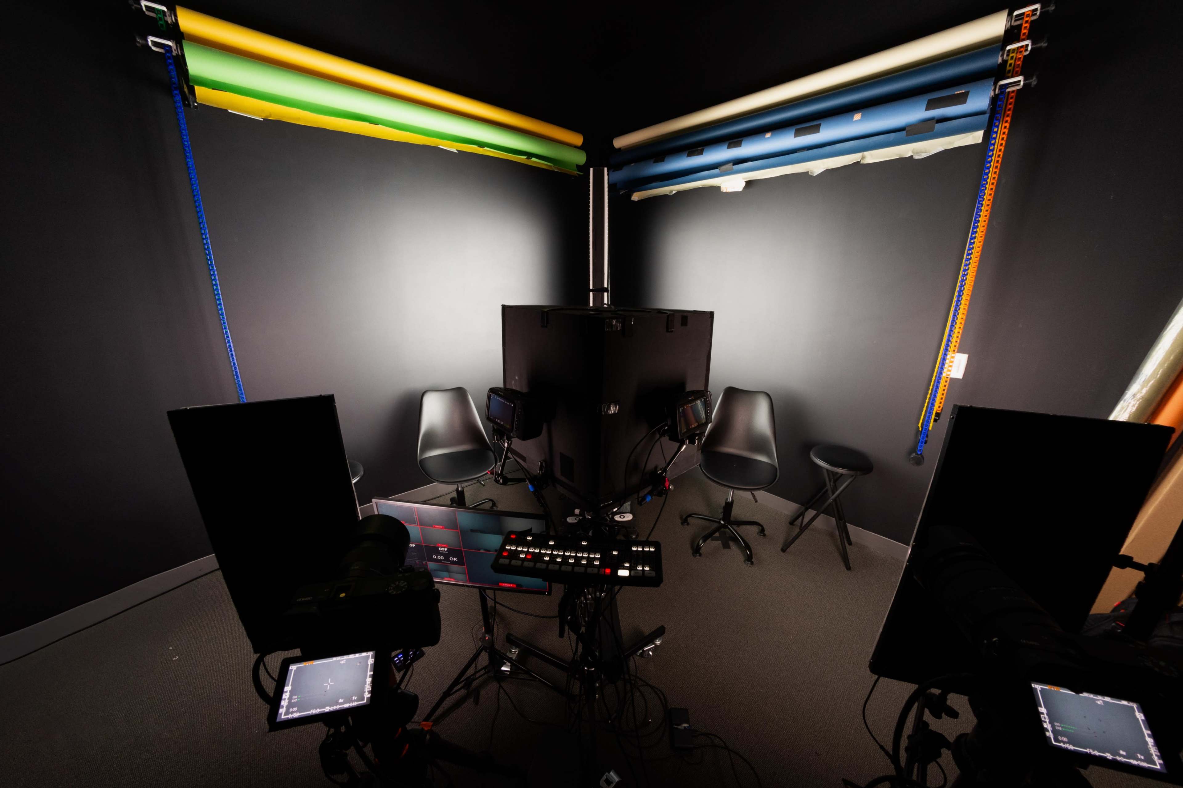 The image shows a photography studio setup with black walls, a central workstation, and backdrop rolls of various colors hanging from the ceiling.
