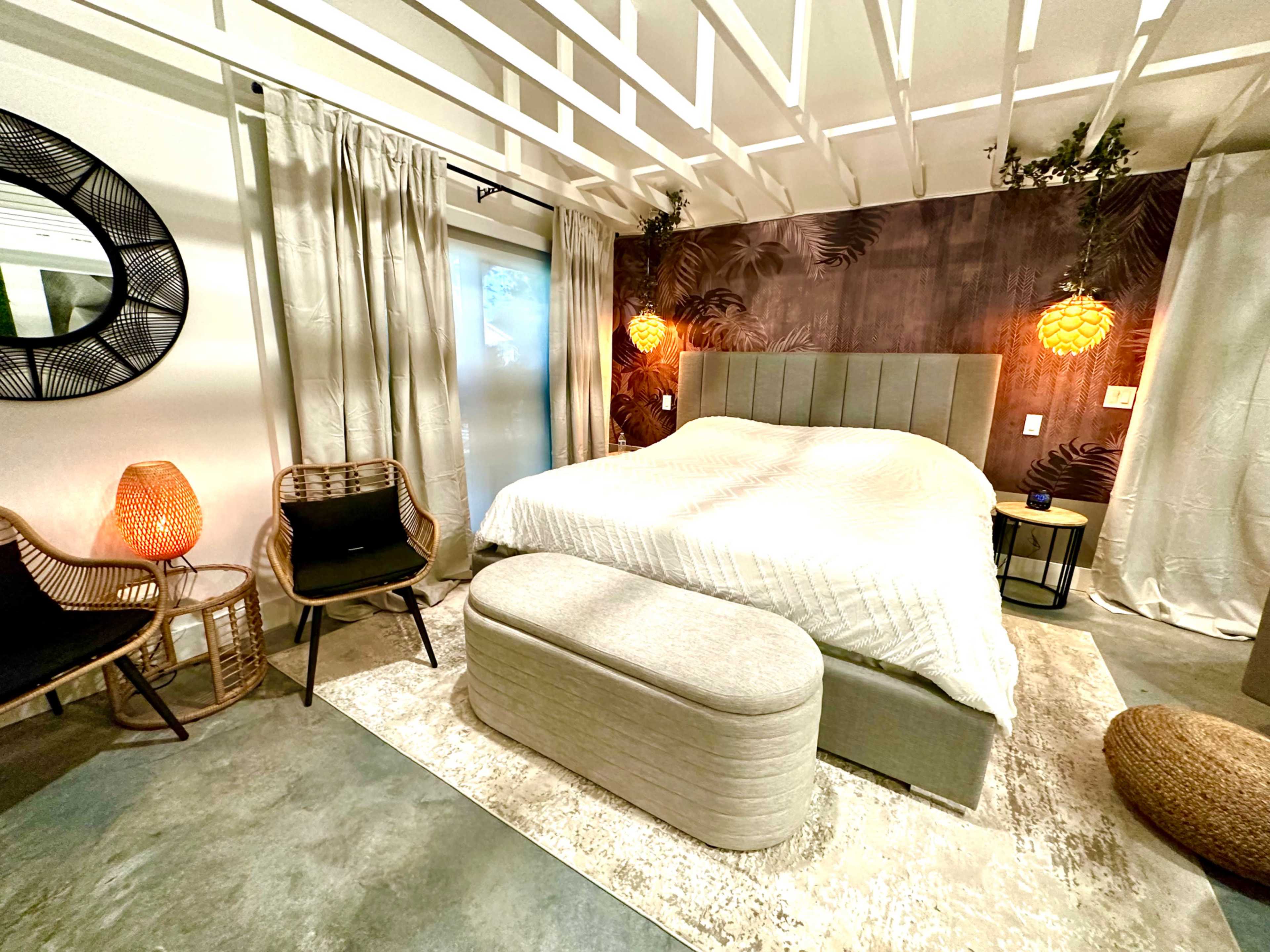 A modern bedroom featuring a large bed with a beige headboard, surrounded by stylish lighting, a circular mirror, and a pair of woven chairs.
