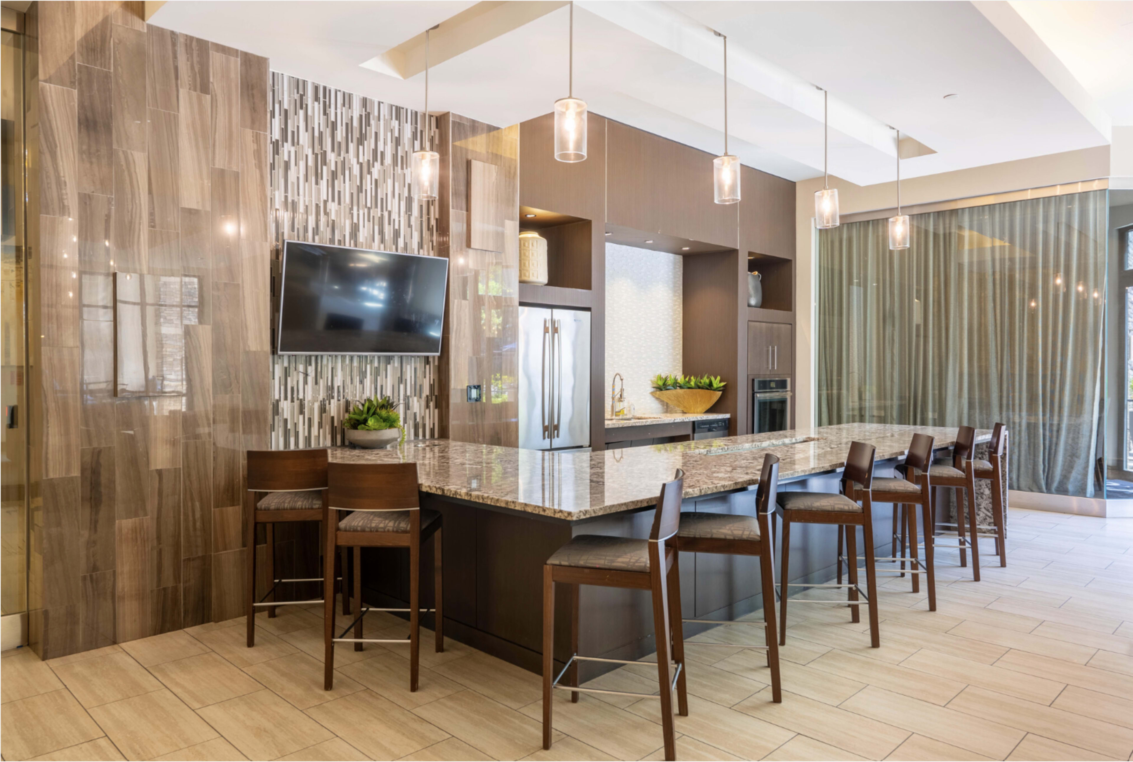 The image shows a modern kitchen with a large island, several bar stools, and pendant lighting, featuring a television mounted on the wall and a stainless steel refrigerator.