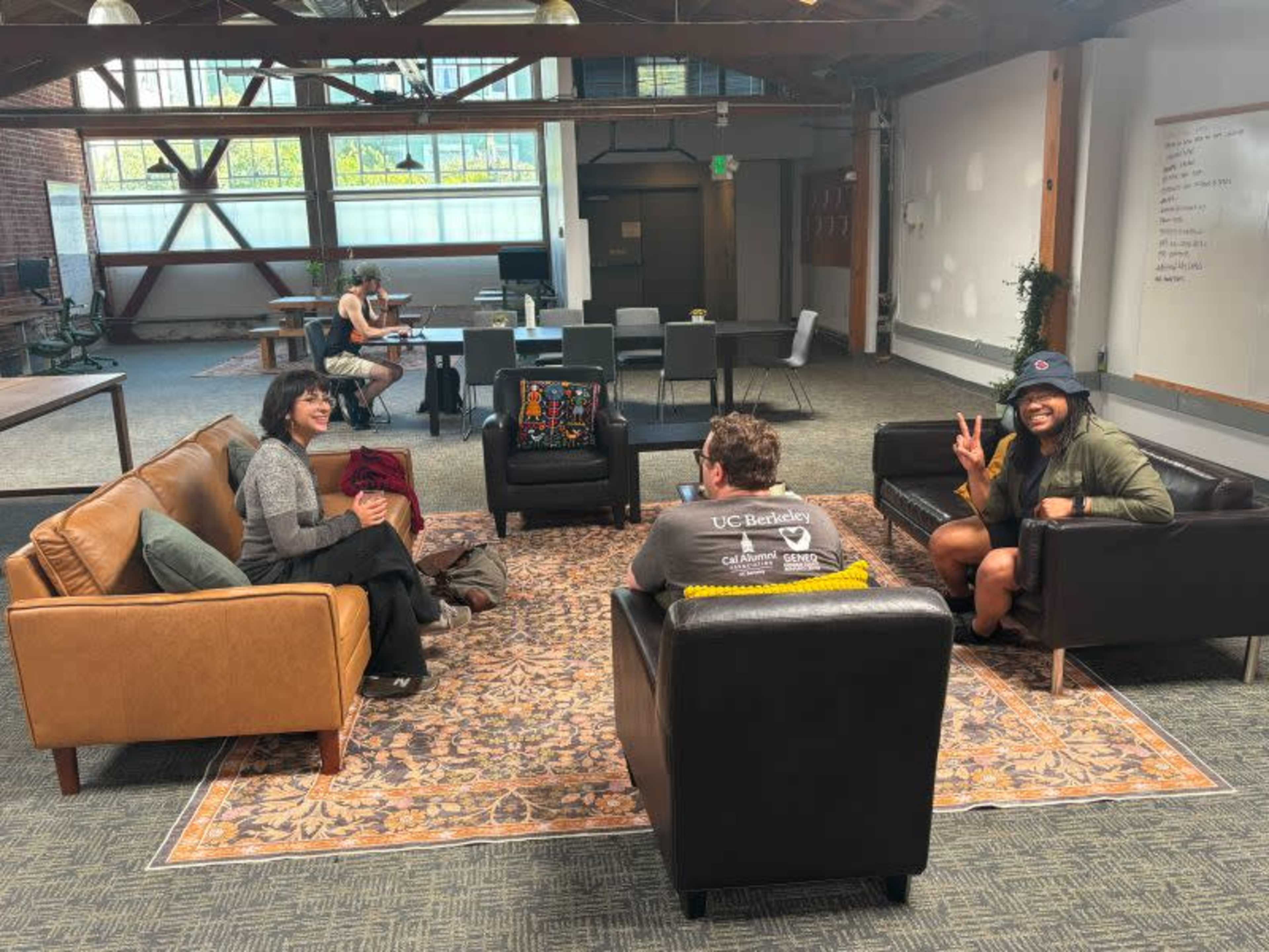 A group of three people are seated in a spacious, modern workspace with sofas and a large area rug, while others are visible in the background engaged in various activities.