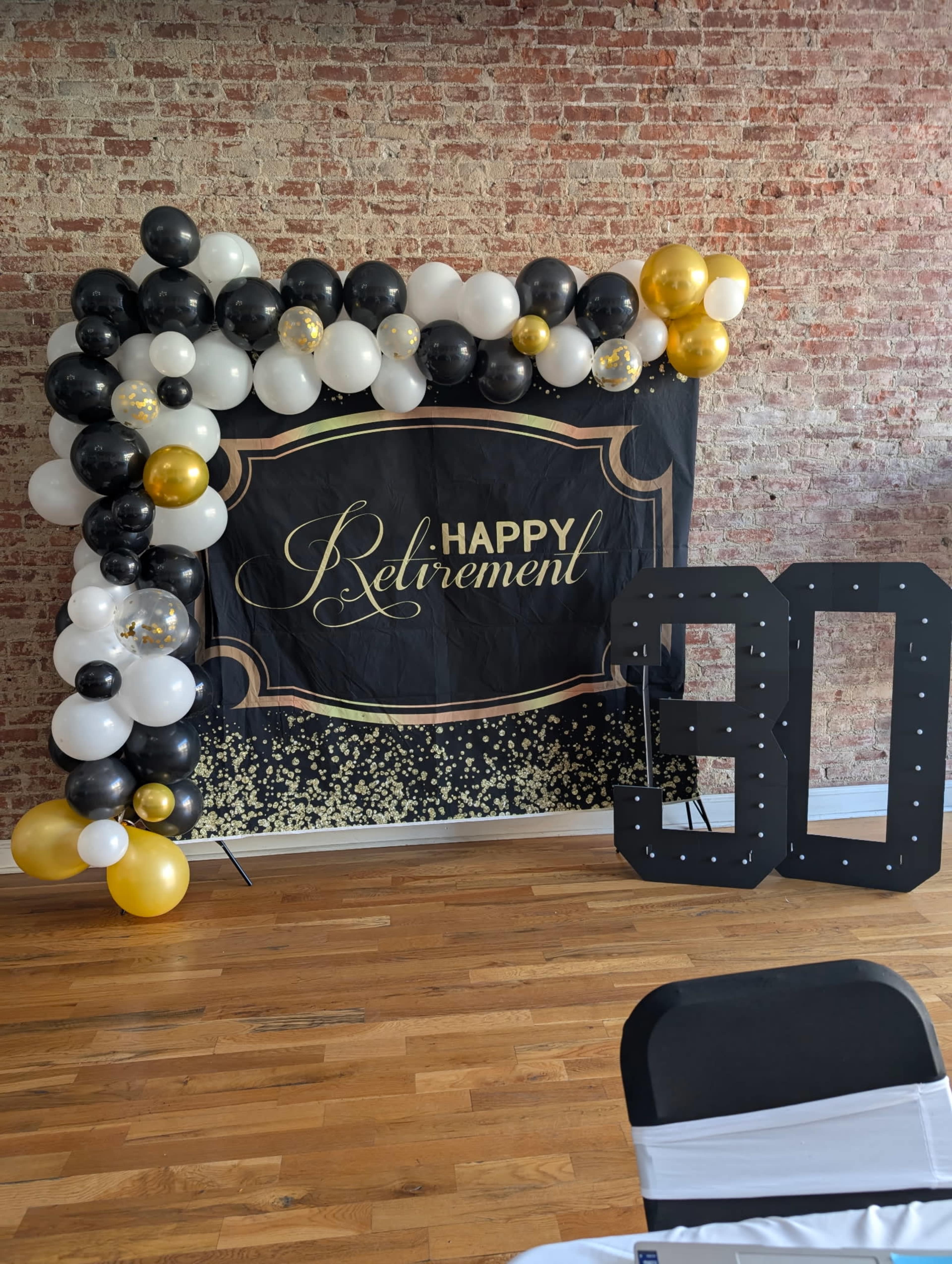 The image shows a decorated backdrop for a retirement celebration featuring black, white, and gold balloons, with a large "30" display beside it.