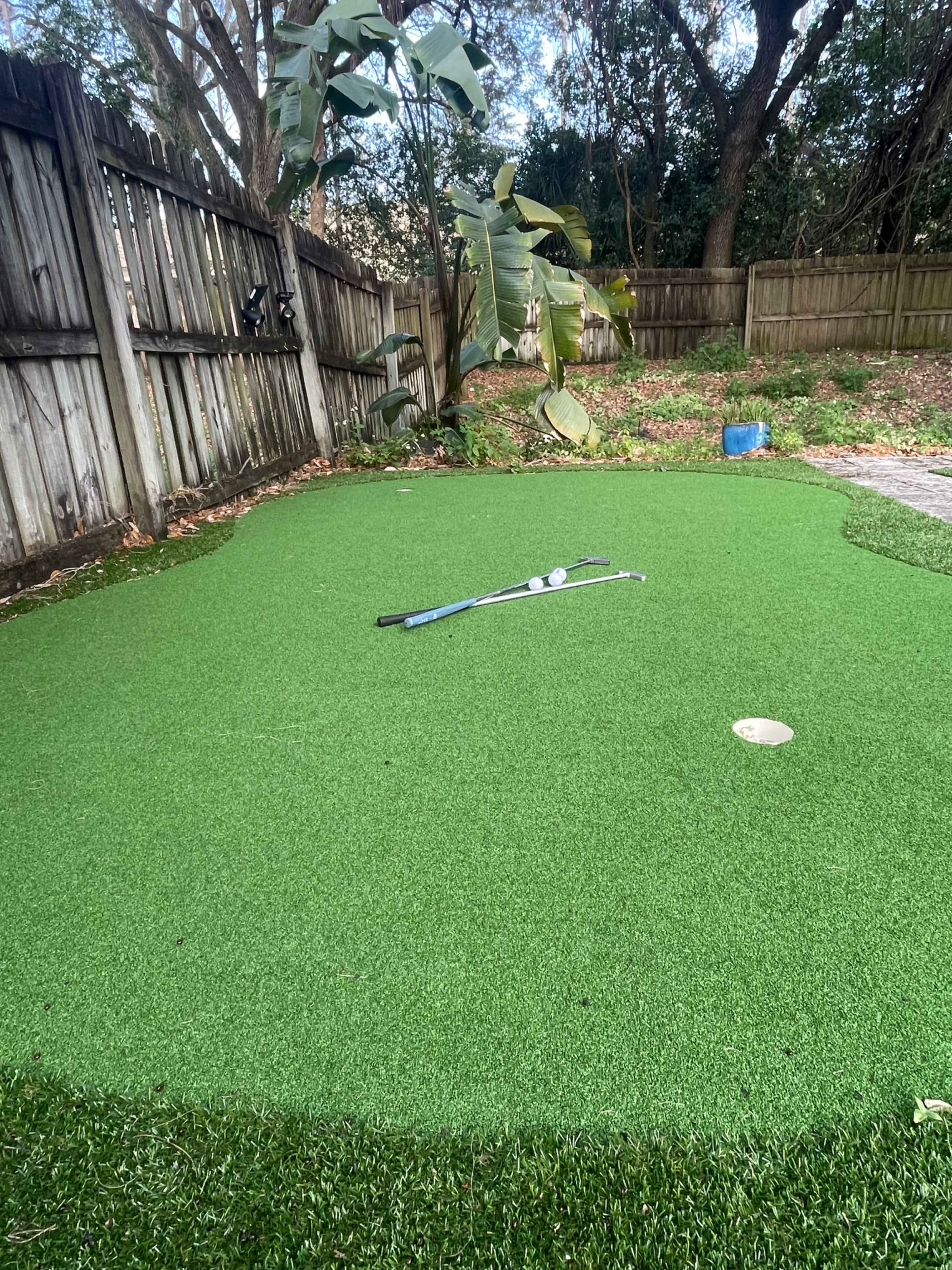 The image shows a small putting green in a backyard, with two golf balls and a putter resting on the artificial turf.