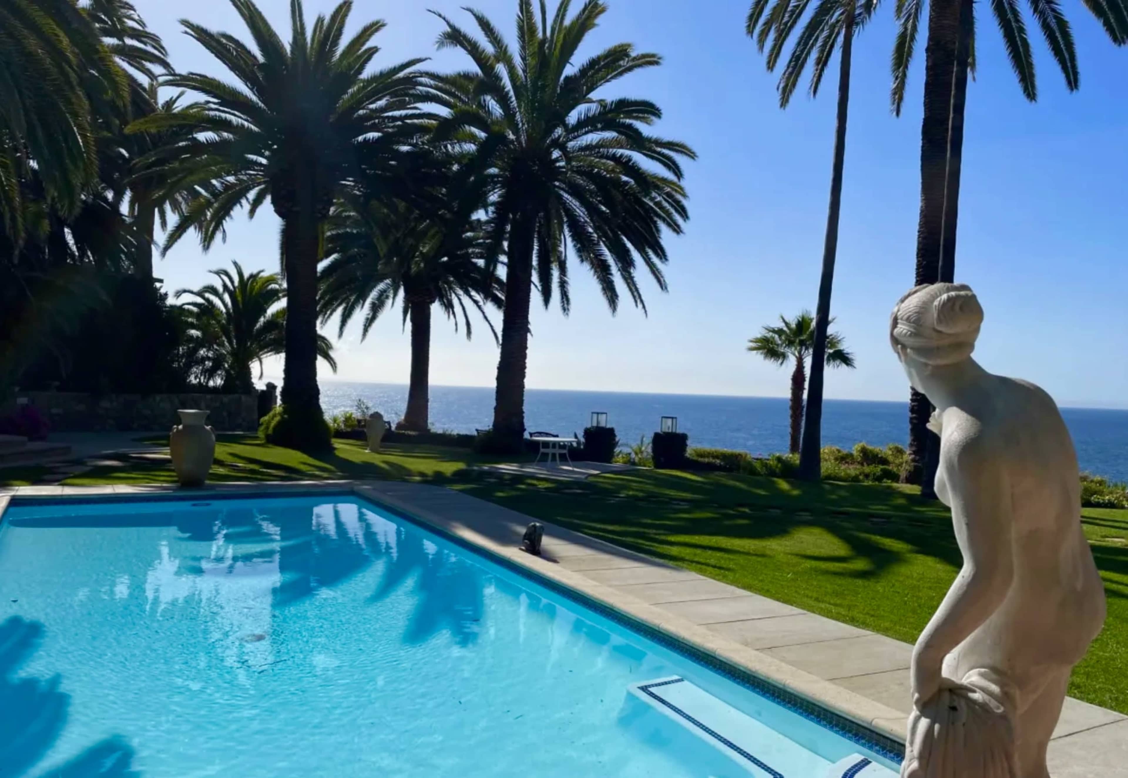 A serene poolside scene with palm trees, a statue, and a view of the ocean in the background.
