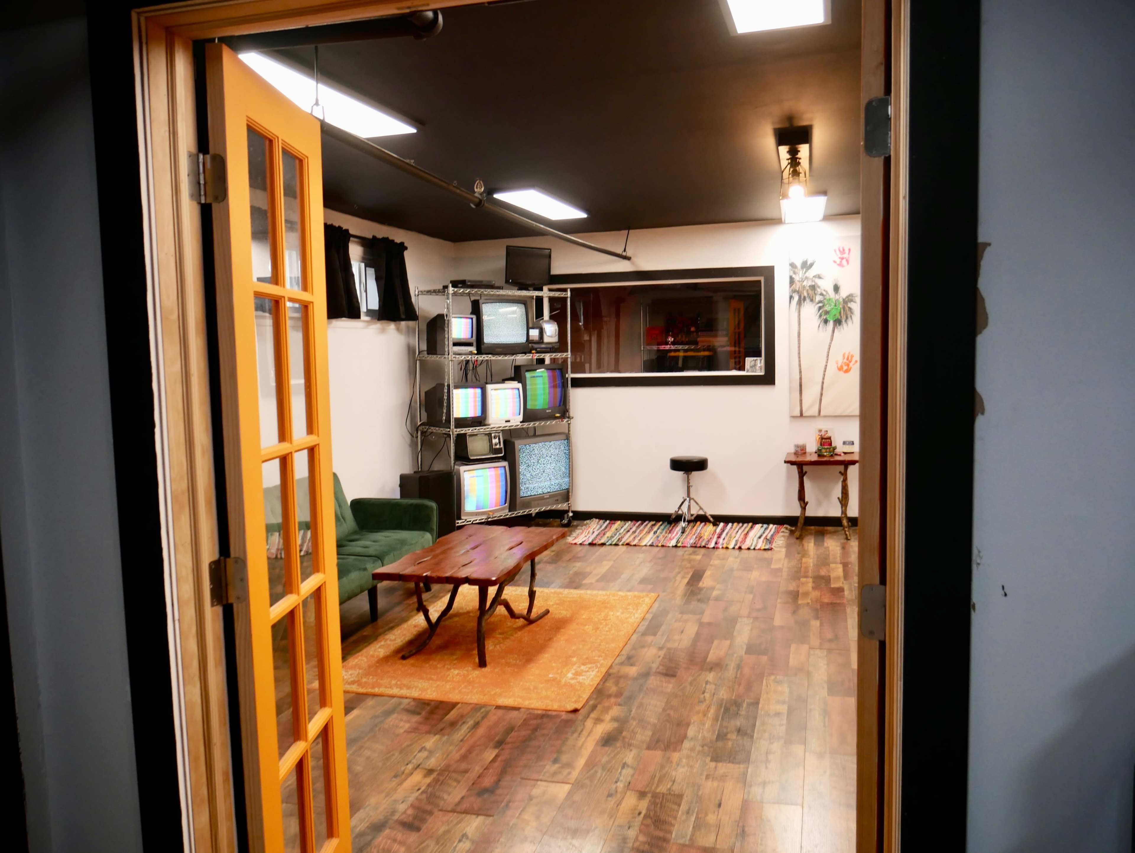 The image shows a room with wooden flooring, a green couch, a wooden coffee table, and several vintage televisions on shelves in the background.
