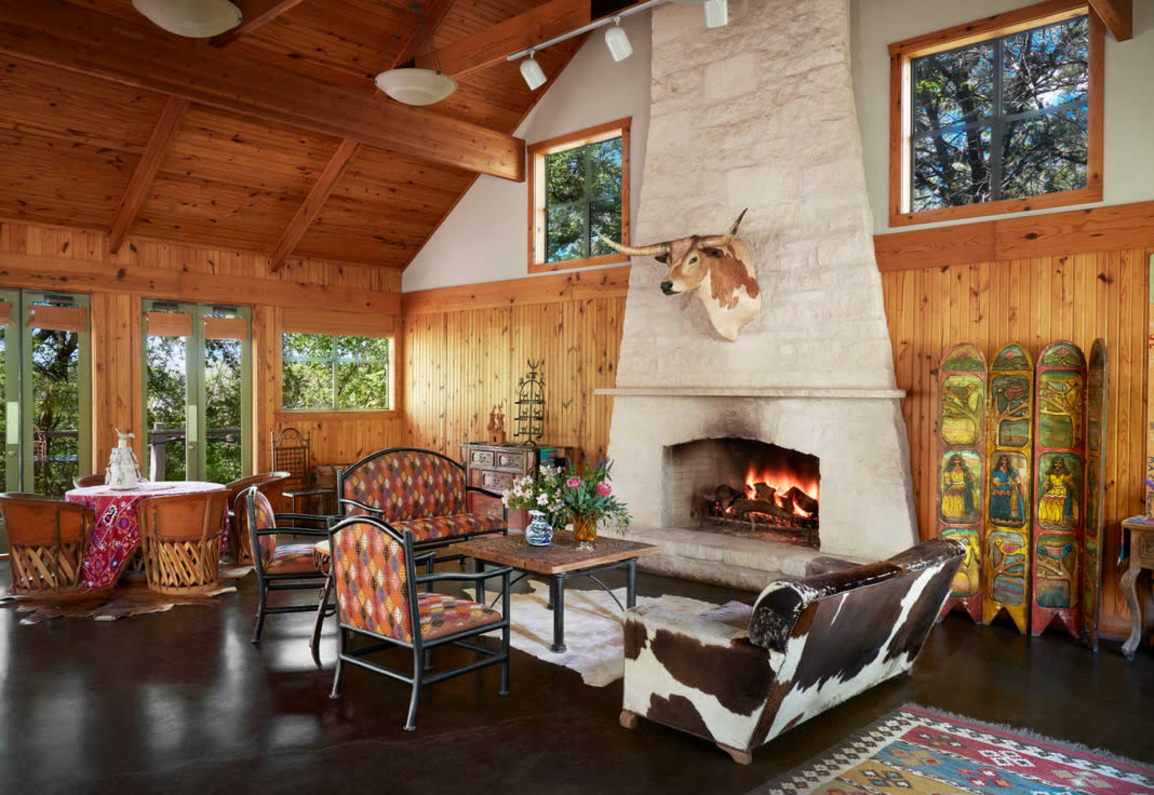 The image depicts a cozy living room adorned with wooden beams, a stone fireplace with a mounted bull's head, and vintage ski gear, surrounded by large windows that offer a view of the outdoors.
