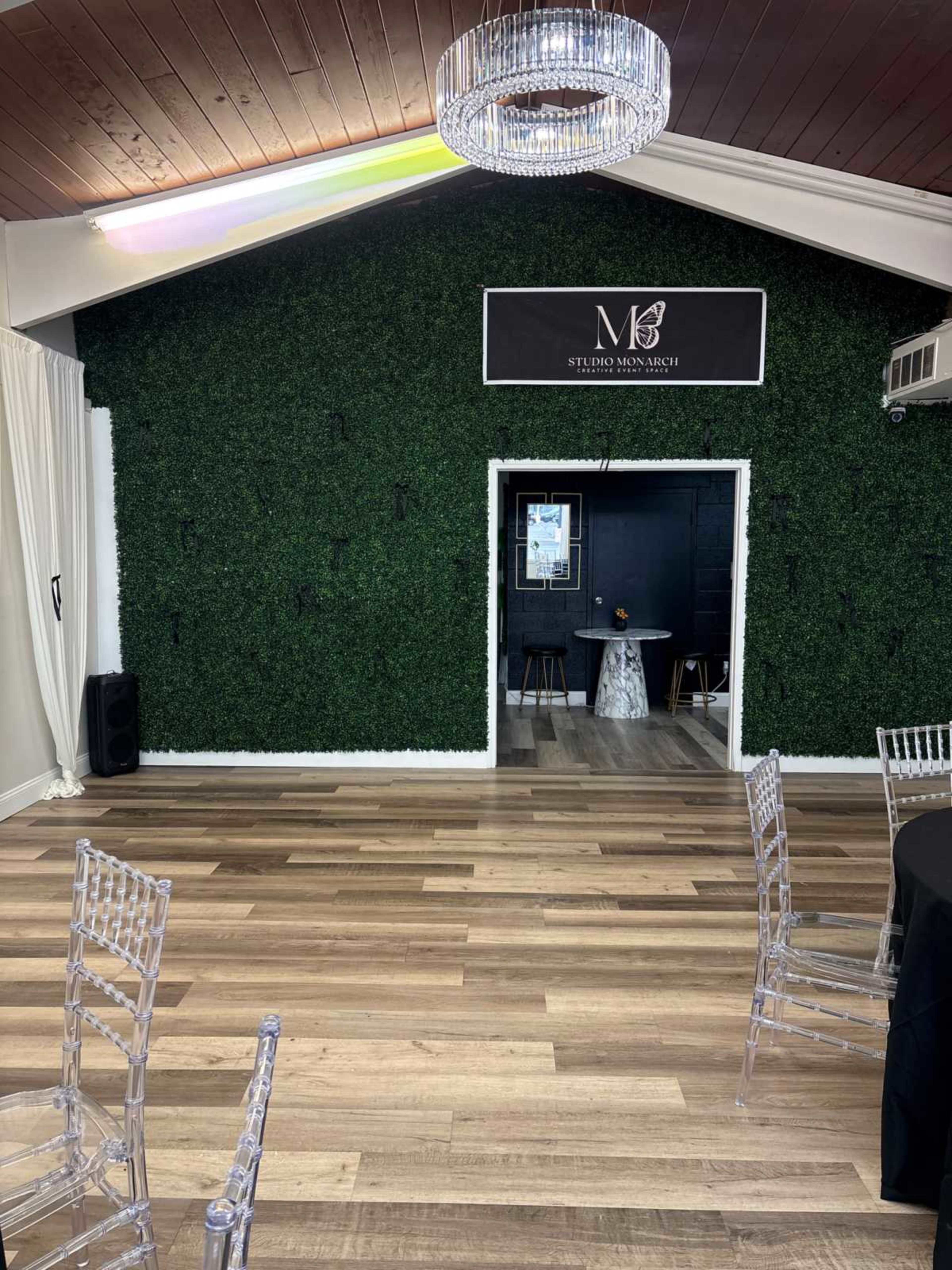 The image shows a room with wooden flooring, a green hedge-like wall, and a black sign displaying "Studio Mon Much" at the center, with transparent chairs arranged around black tables.