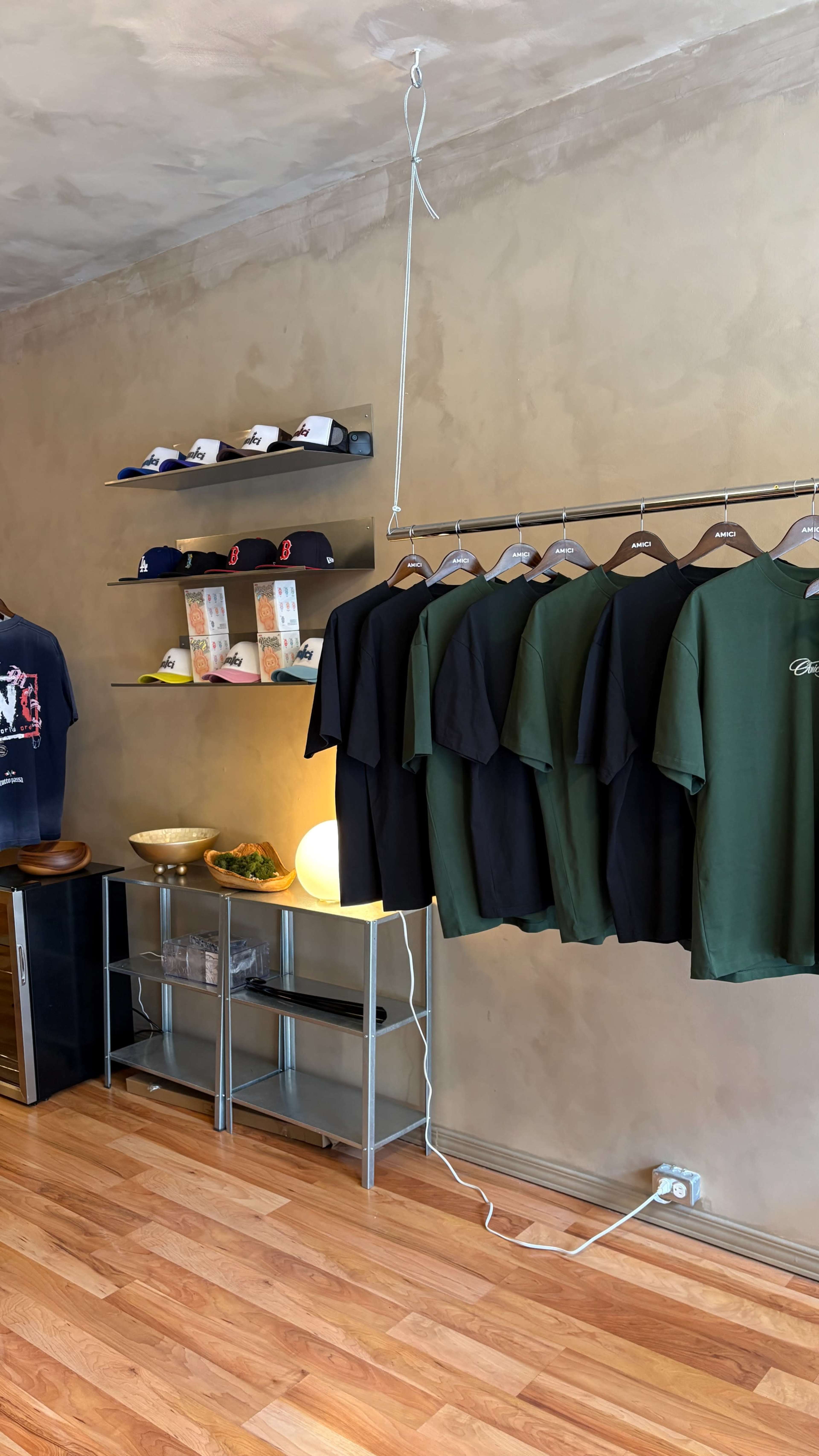 The image shows a clothing store interior featuring shelves with caps and a row of T-shirts hanging on a rack against a textured wall.