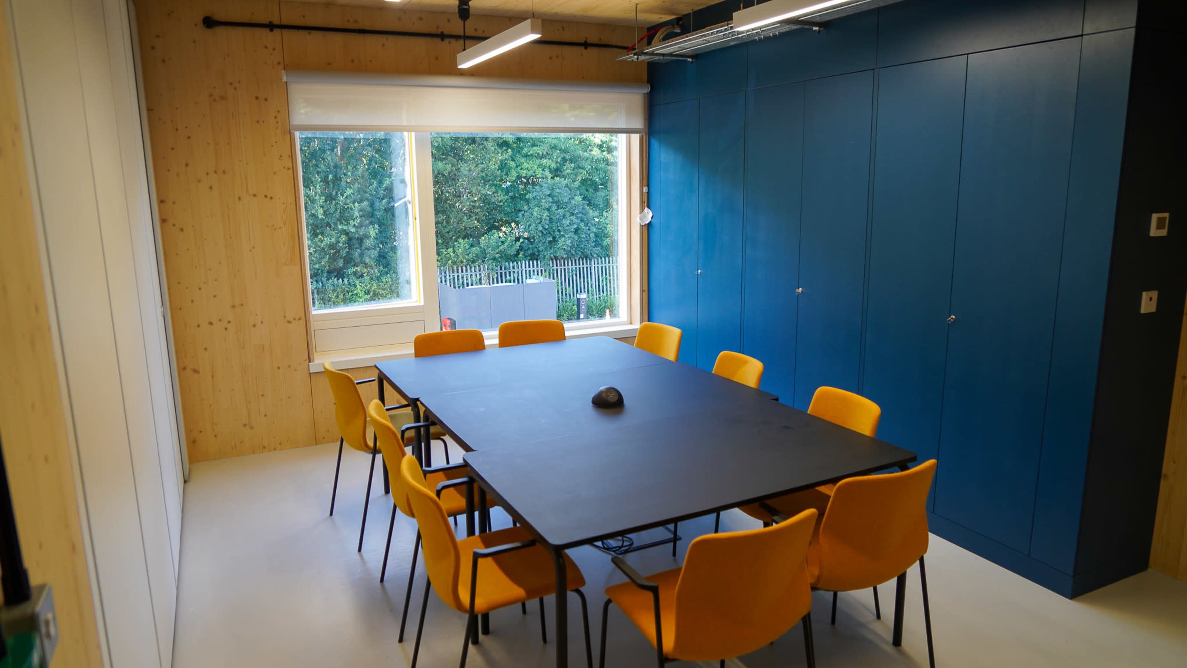 A conference room features a large black table surrounded by orange chairs, with a window overlooking greenery.