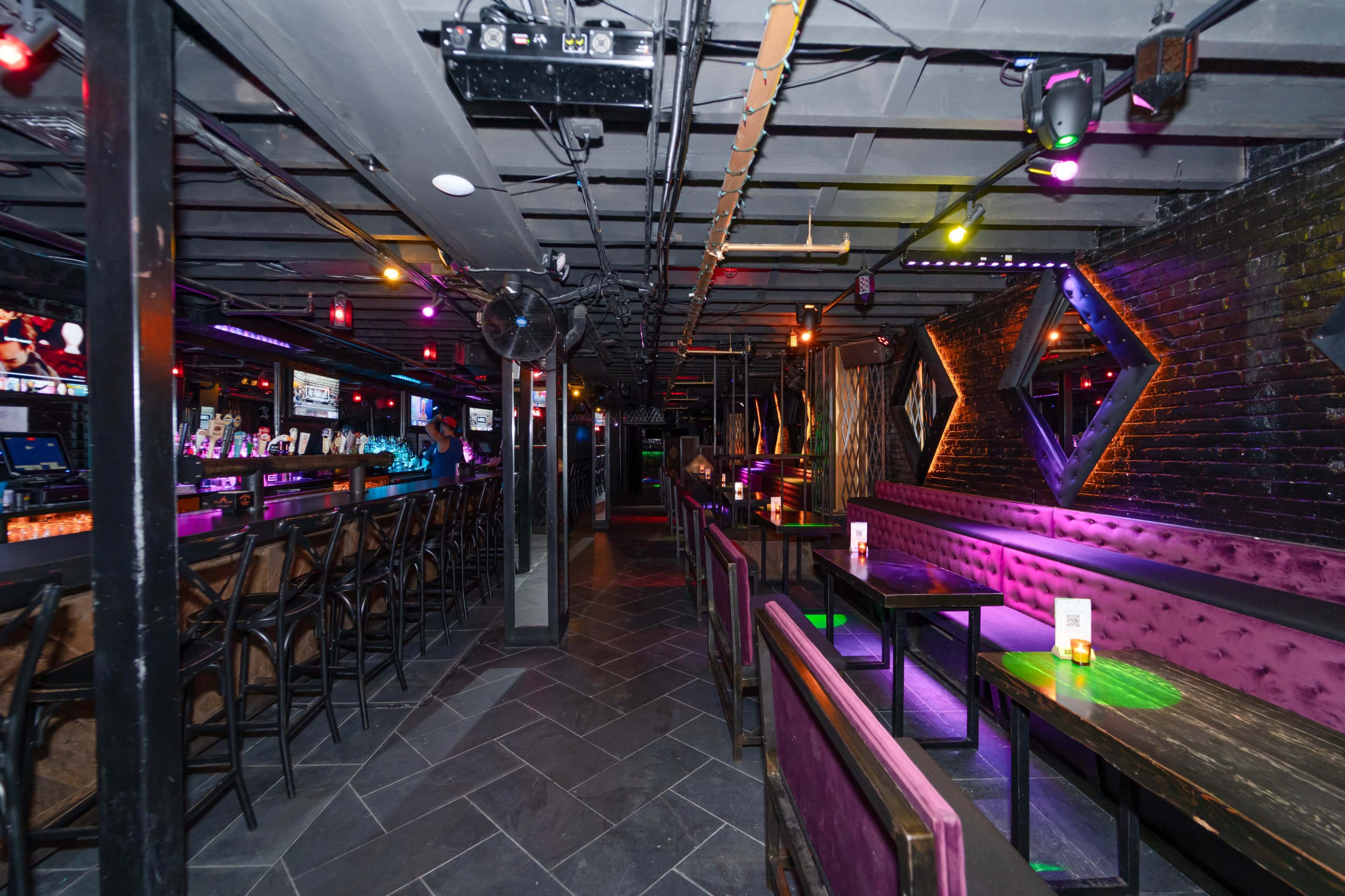 A dimly lit bar with a long counter, high stools, and seating areas featuring purple lighting and dark walls.