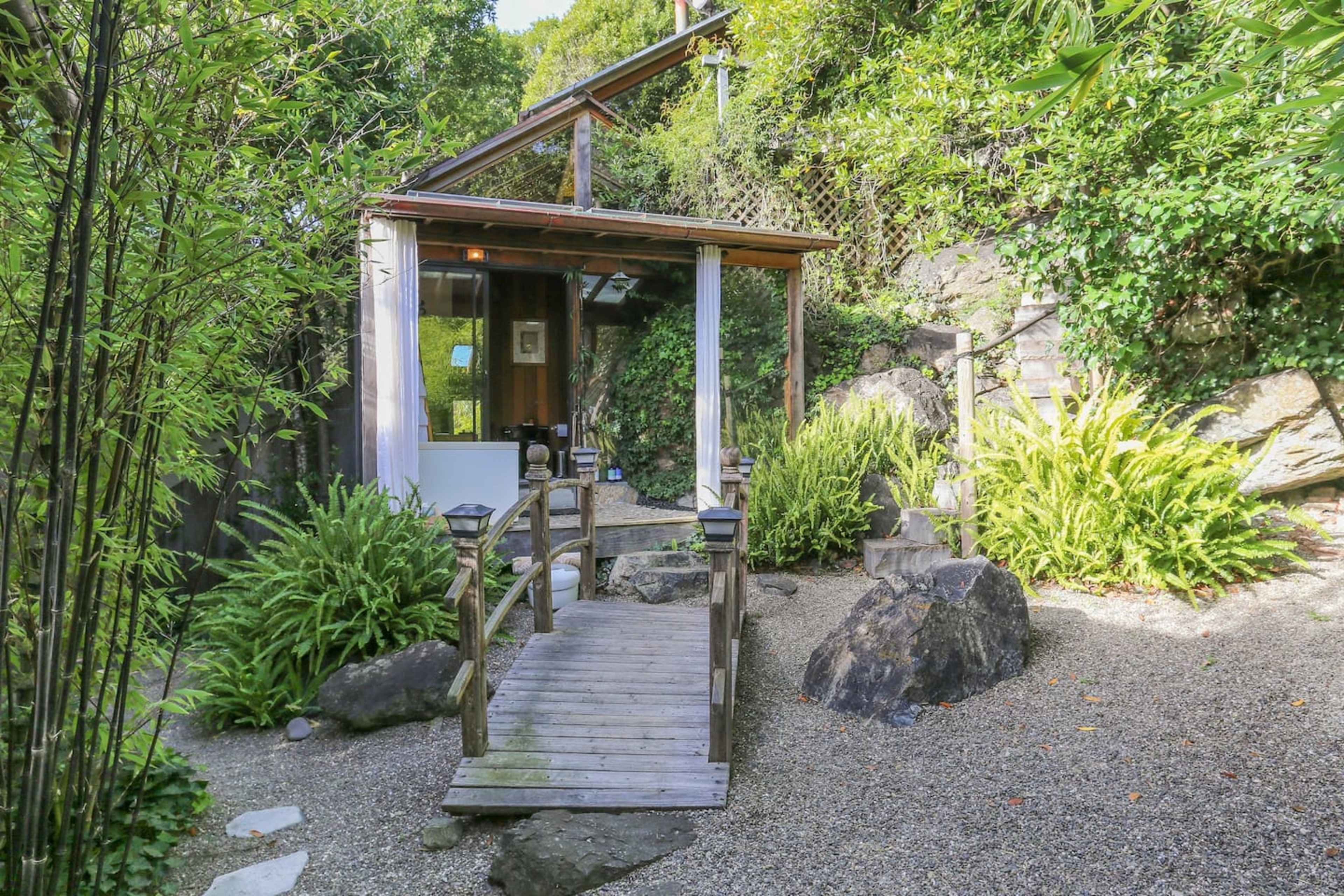 A wooden bridge leads to a small building surrounded by rocks and lush greenery in a landscaped garden.