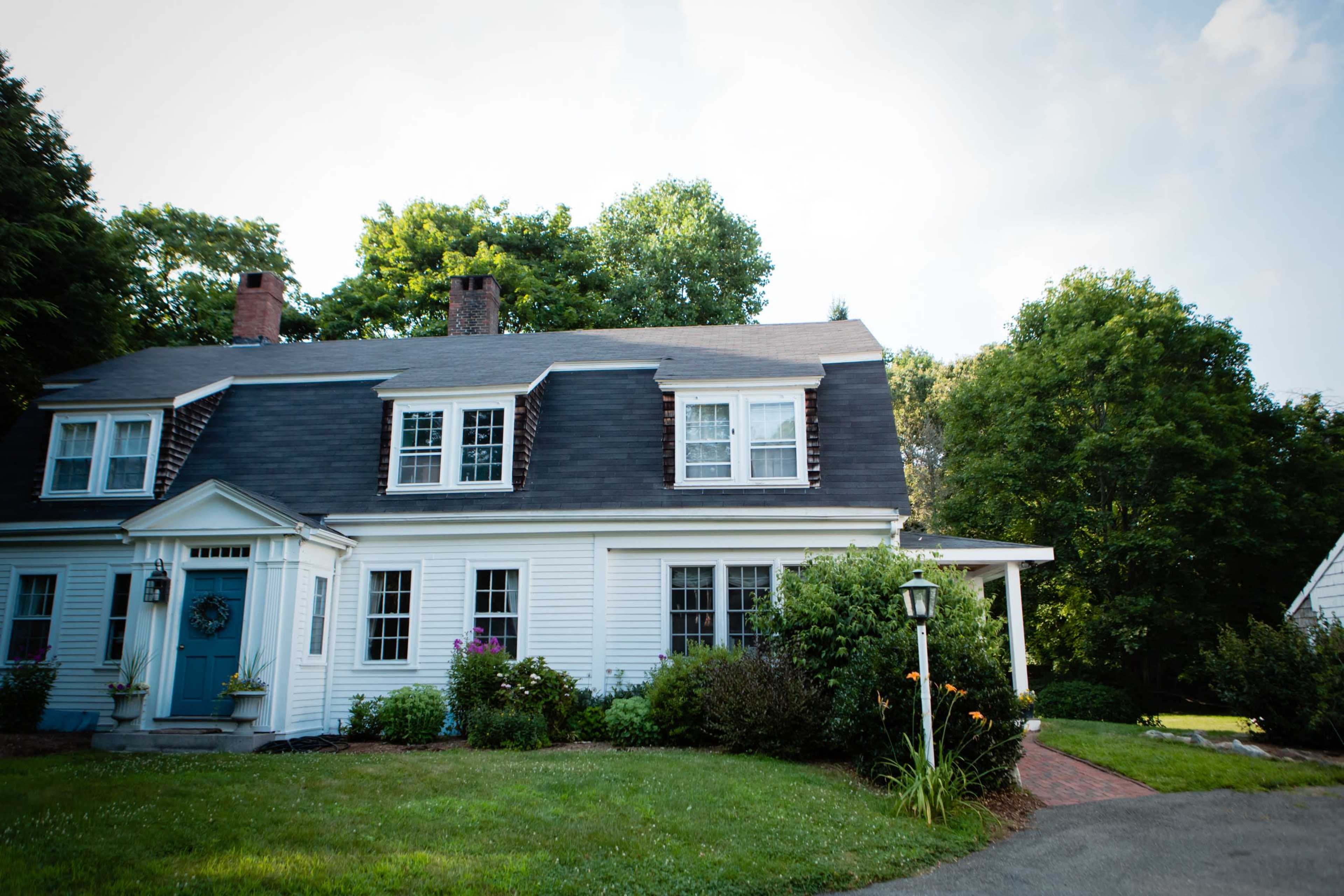A two-story white house with a gray roof and a blue front door is surrounded by green trees and shrubs.