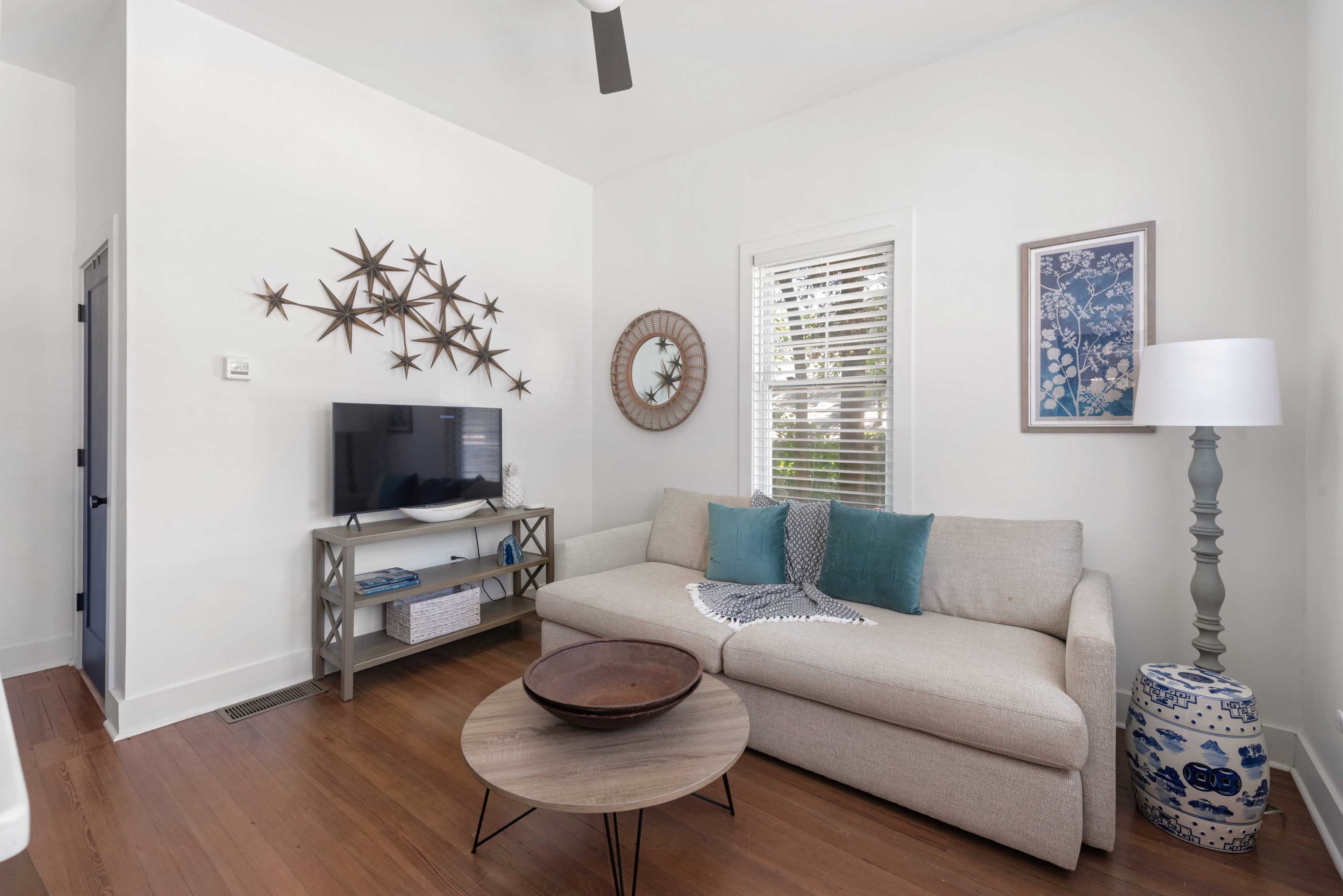 The image shows a small living room with a beige sofa, a round wooden coffee table, a television on a stand, and decorative wall art.