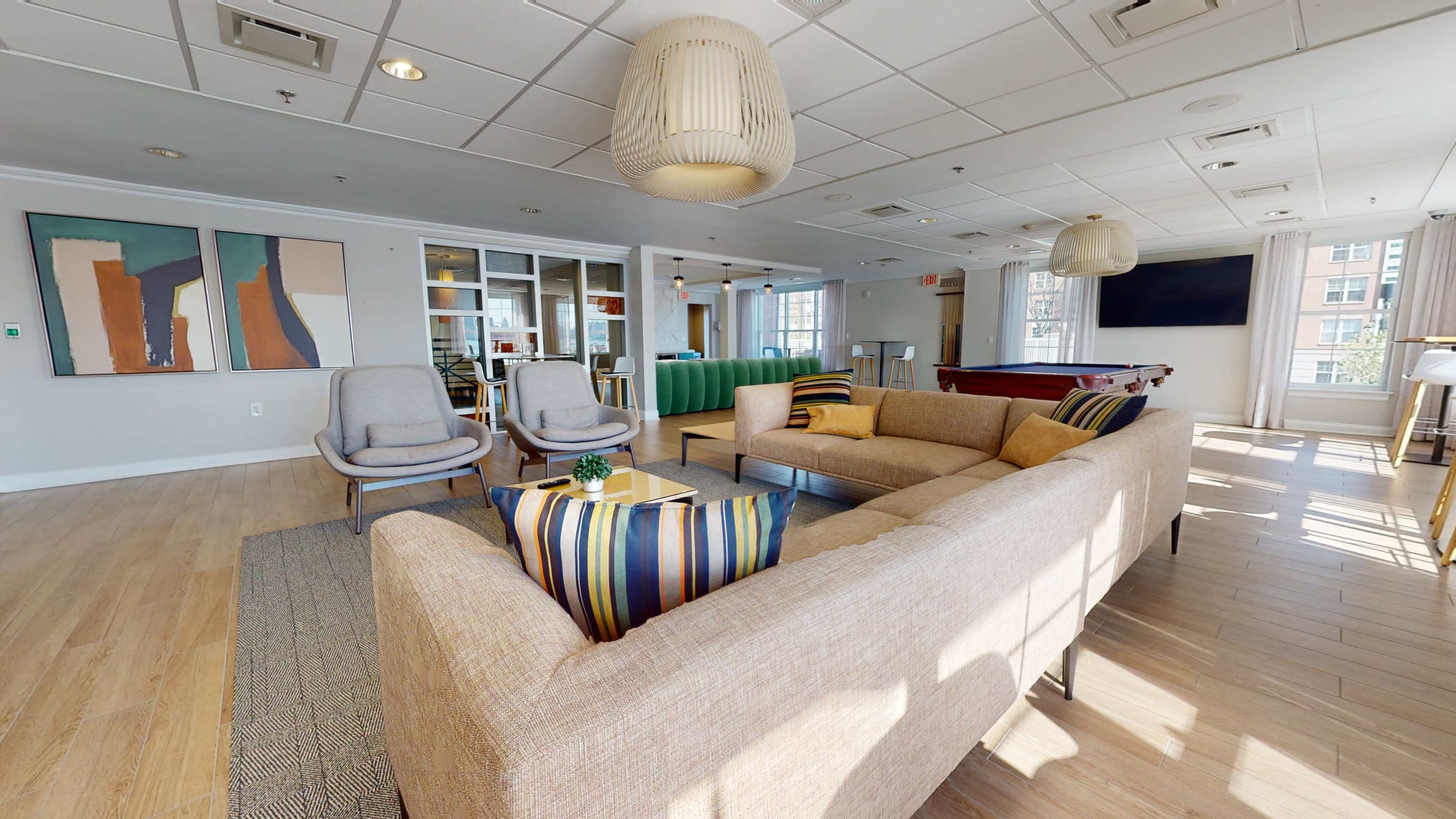 A modern lounge area with a beige sectional sofa, two armchairs, and a pool table in a spacious room filled with natural light.
