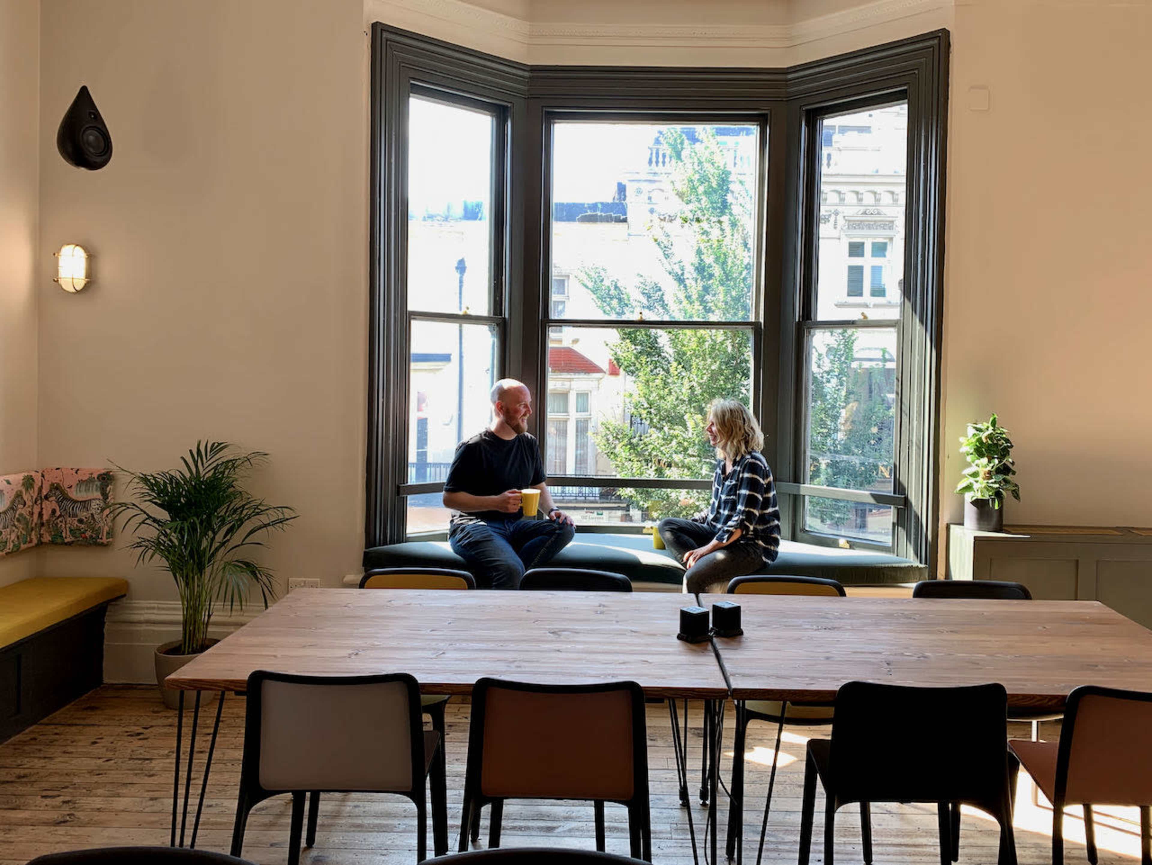 A man and a child sit on a window ledge conversing, with a large table and plants in the room.