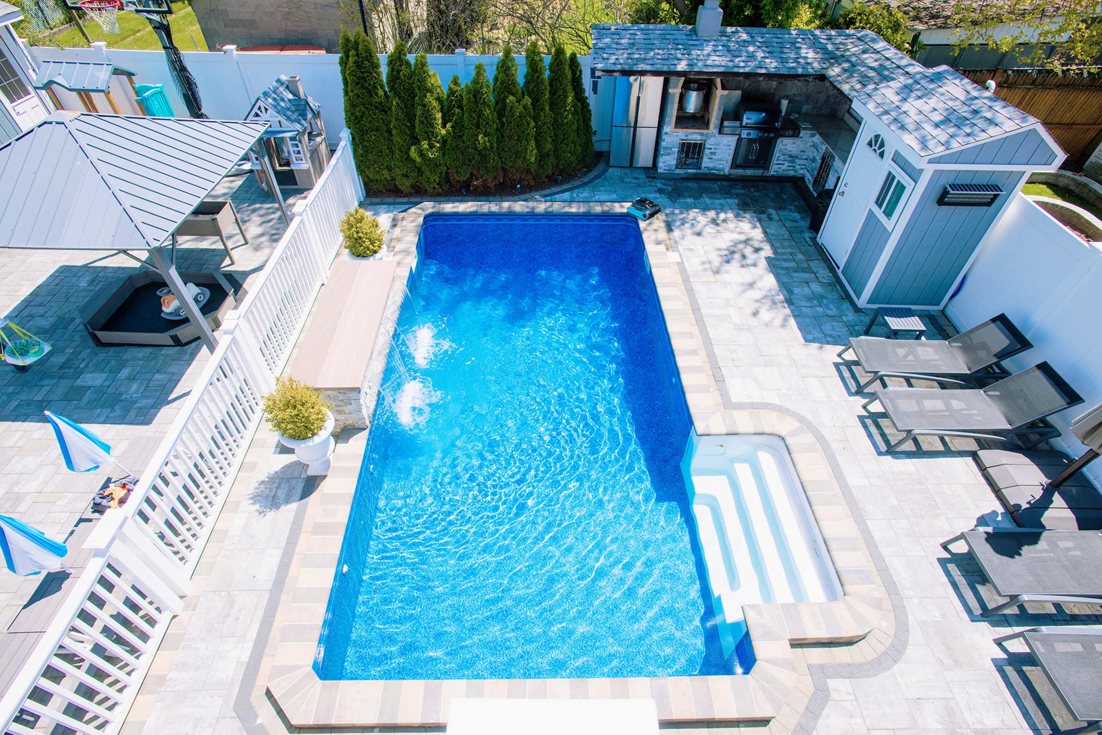 The image shows a rectangular swimming pool surrounded by stone tiles, lounging chairs, and a small spa area, with a covered outdoor kitchen and a shaded seating area in the background.