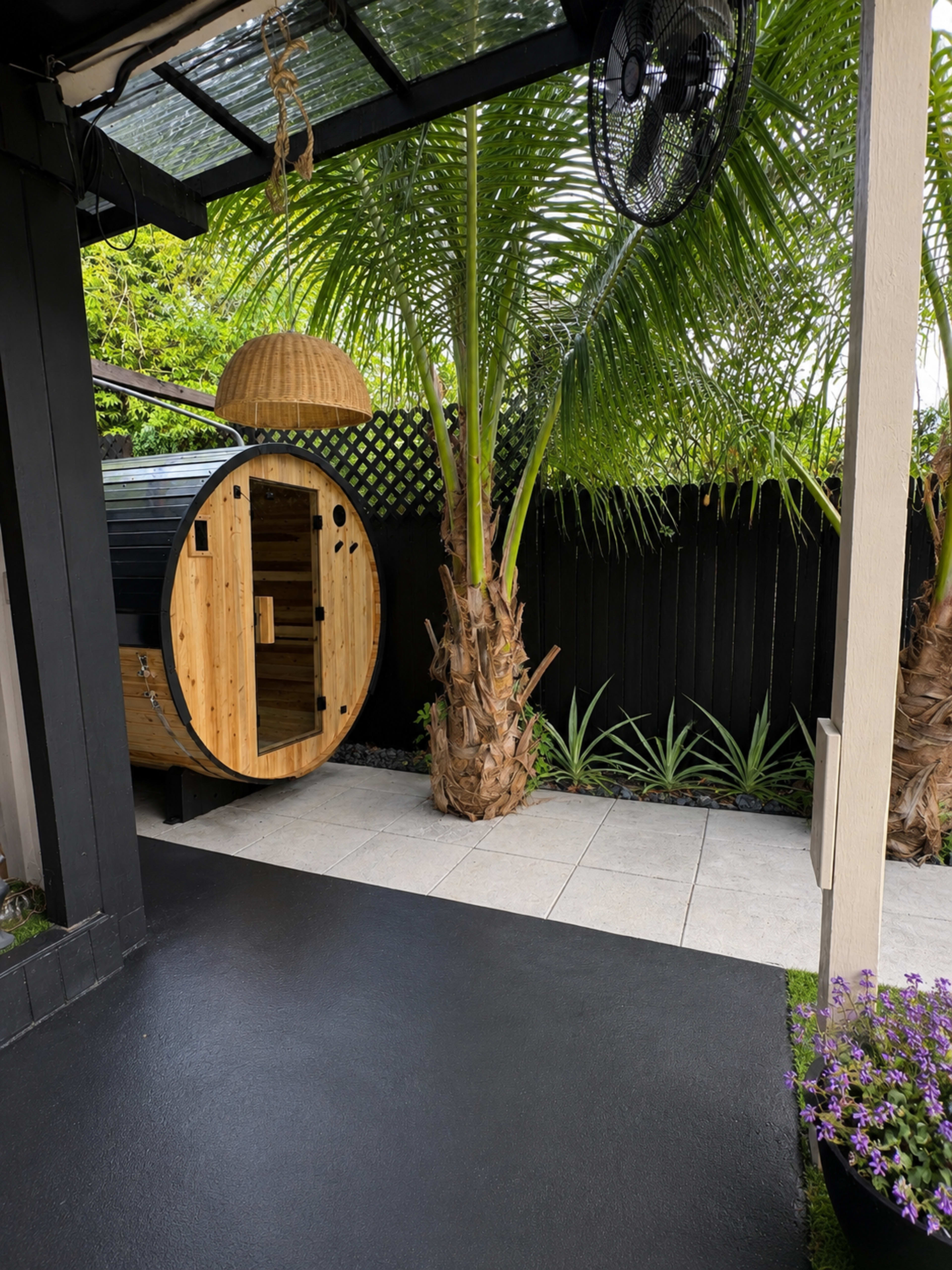 The image shows a backyard area featuring a circular wooden sauna, a tall palm tree, and paved pathways surrounded by greenery.