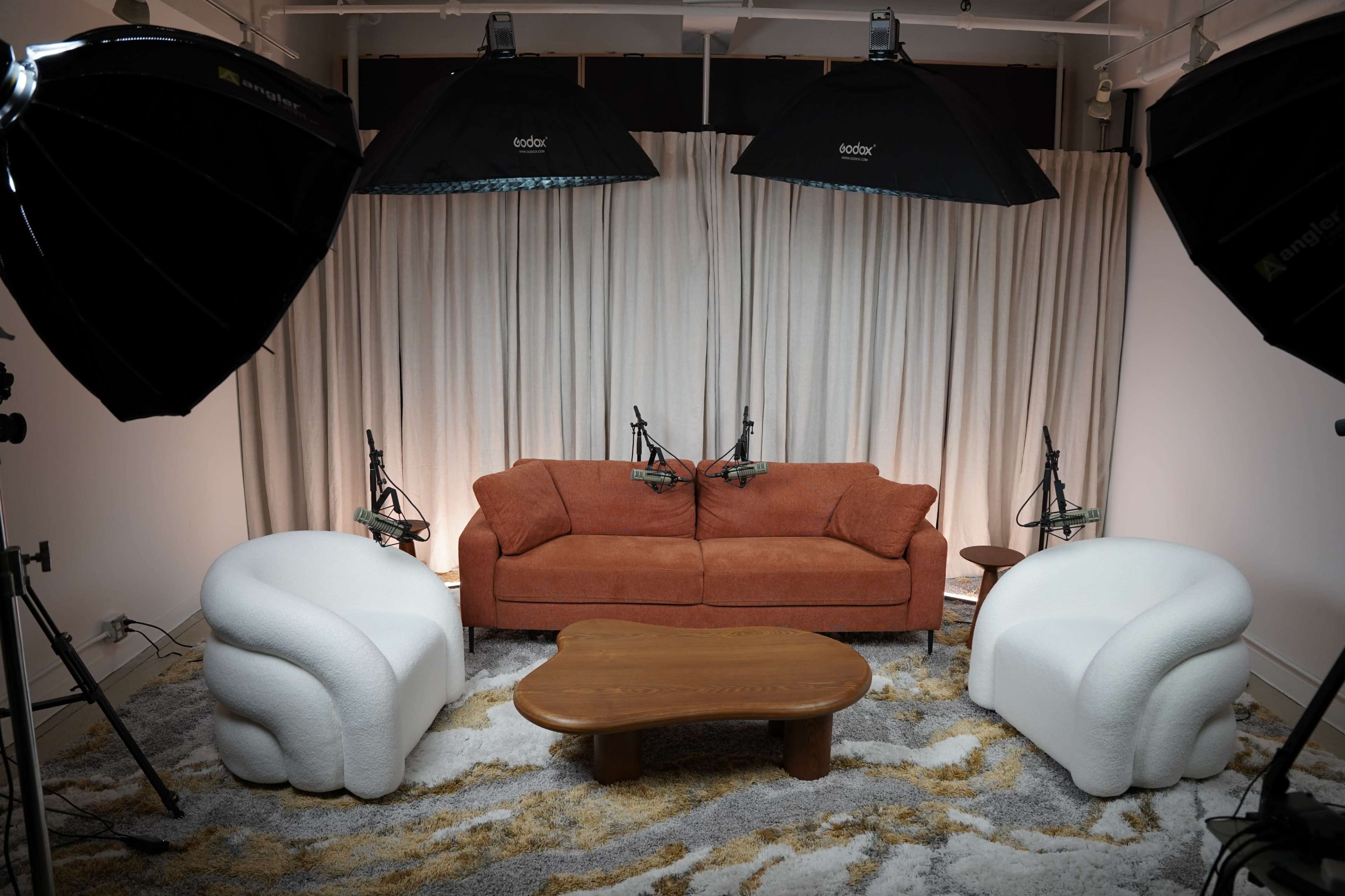 The image shows a cozy living room setup with a brown sofa and two white accent chairs, surrounded by studio lighting and a wooden coffee table on a patterned rug.