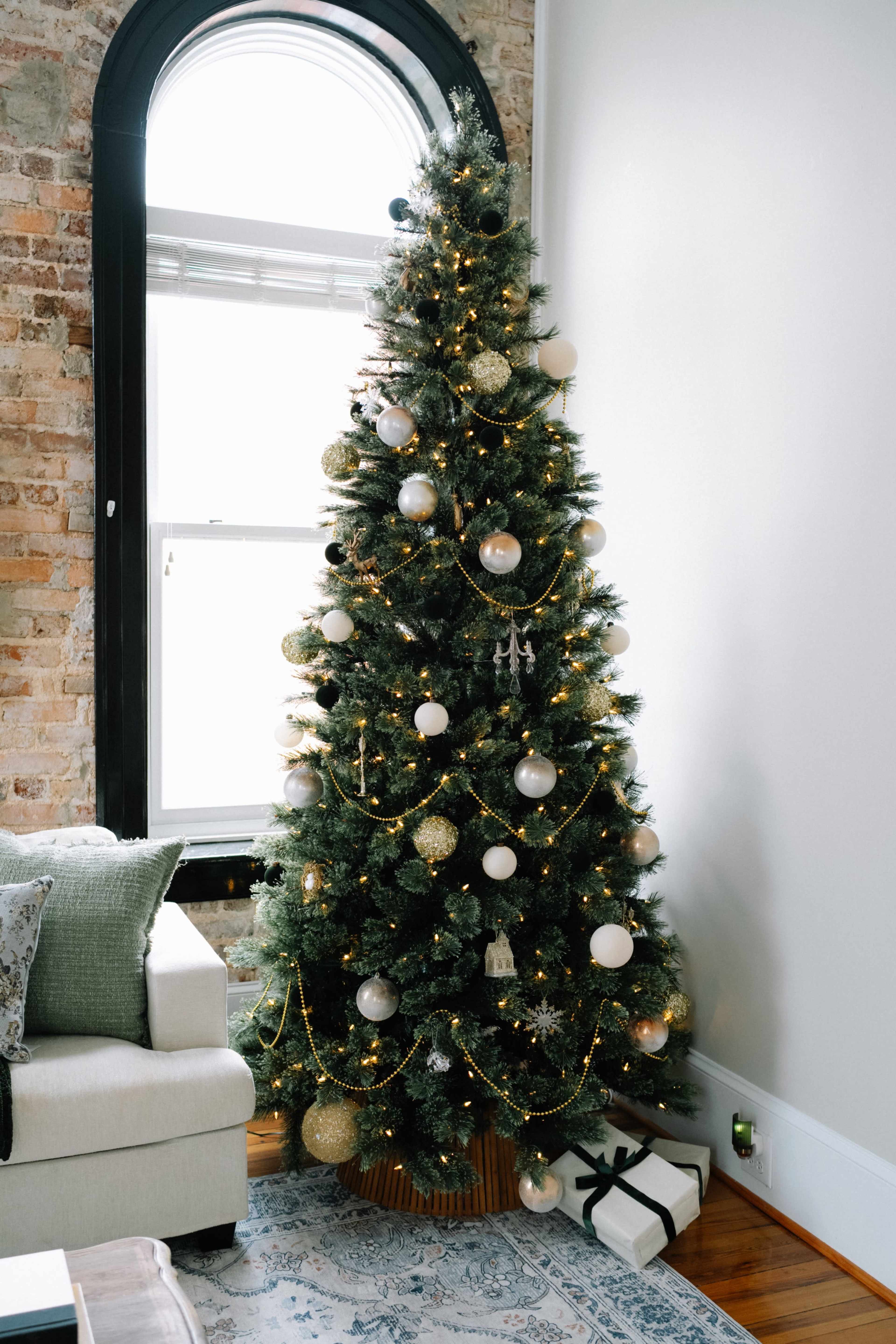 A decorated Christmas tree with silver and gold ornaments stands beside a window in a room featuring exposed brick walls and a neutral-colored sofa.