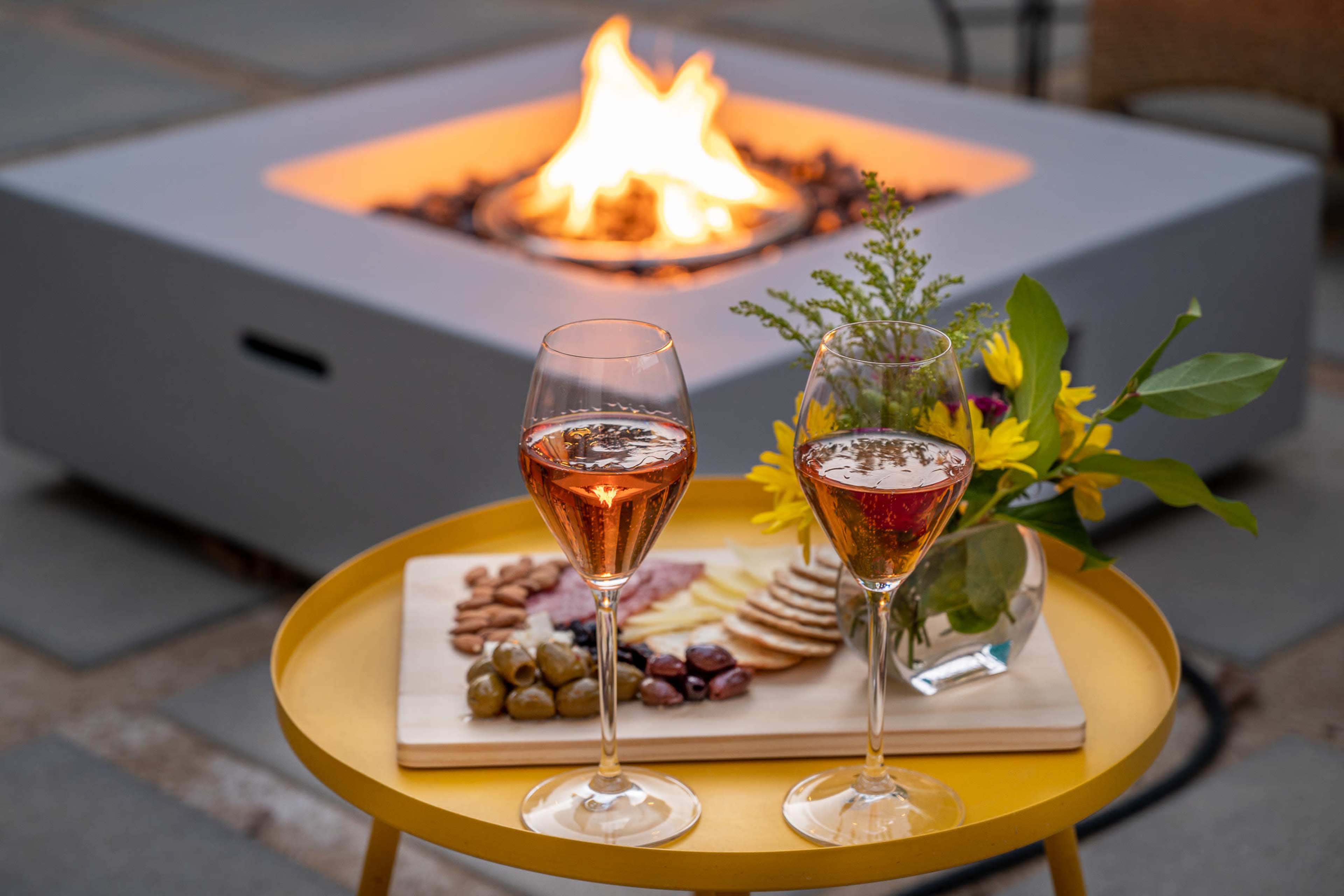Two glasses of rosé wine sit on a yellow table beside a platter of snacks, with a fire pit glowing in the background.