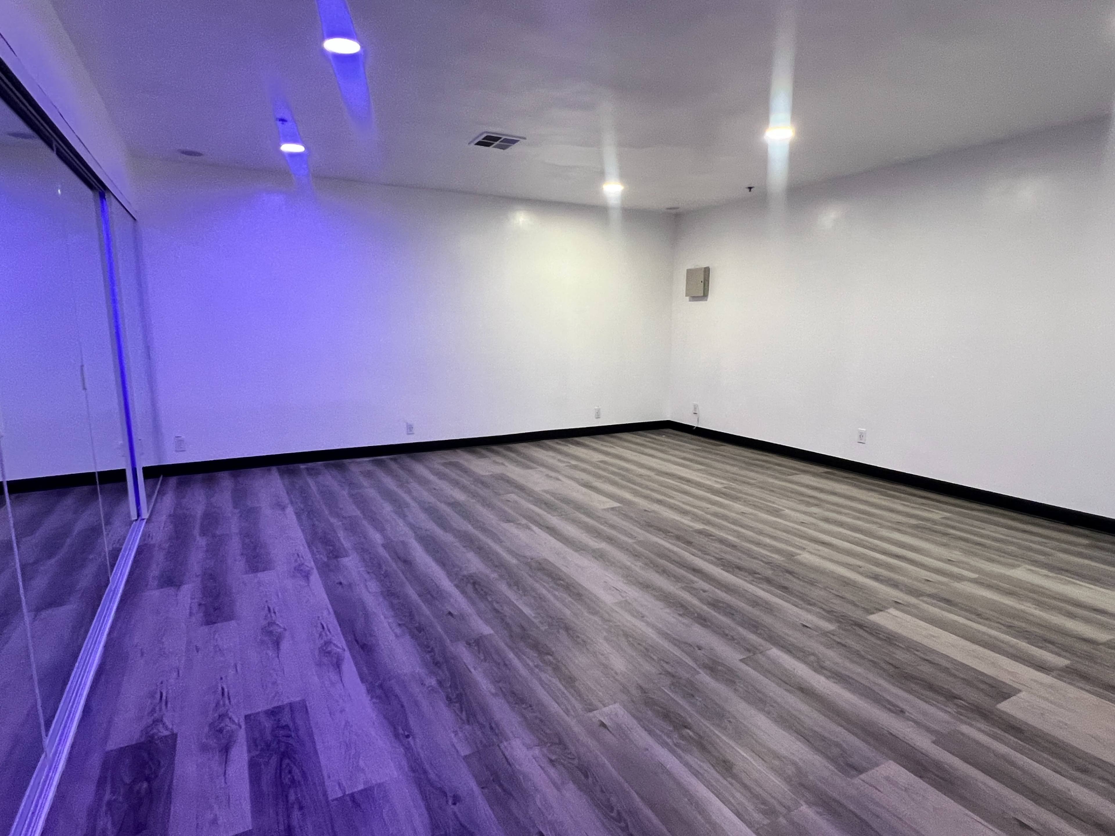 The image shows an empty room with wooden flooring and white walls, featuring recessed lighting and a large mirrored wall.