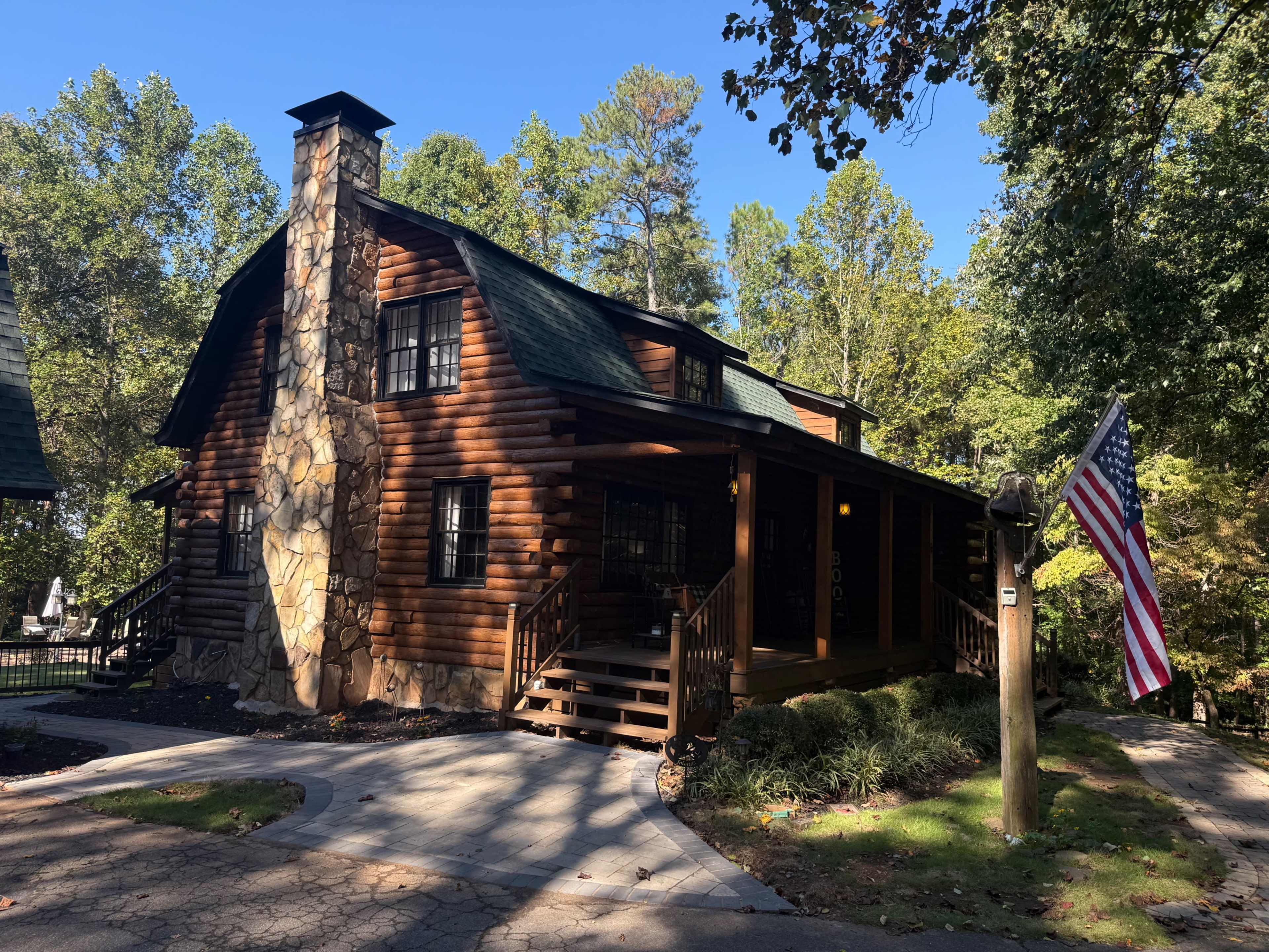 A wooden log cabin with a stone chimney and an American flag stands among trees on a sunny day.