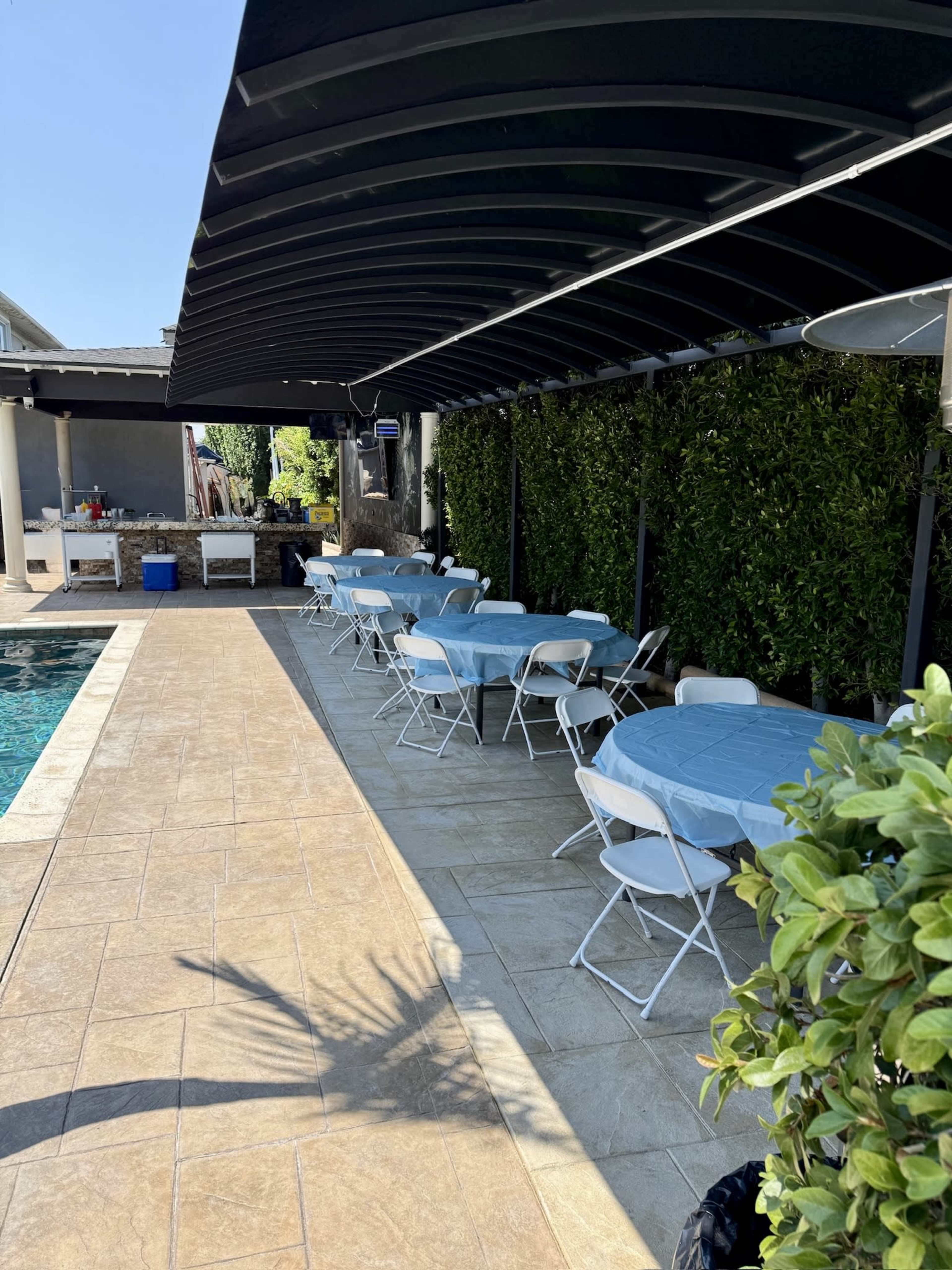 A collection of tables covered with light blue tablecloths is arranged next to a swimming pool under a shaded structure.