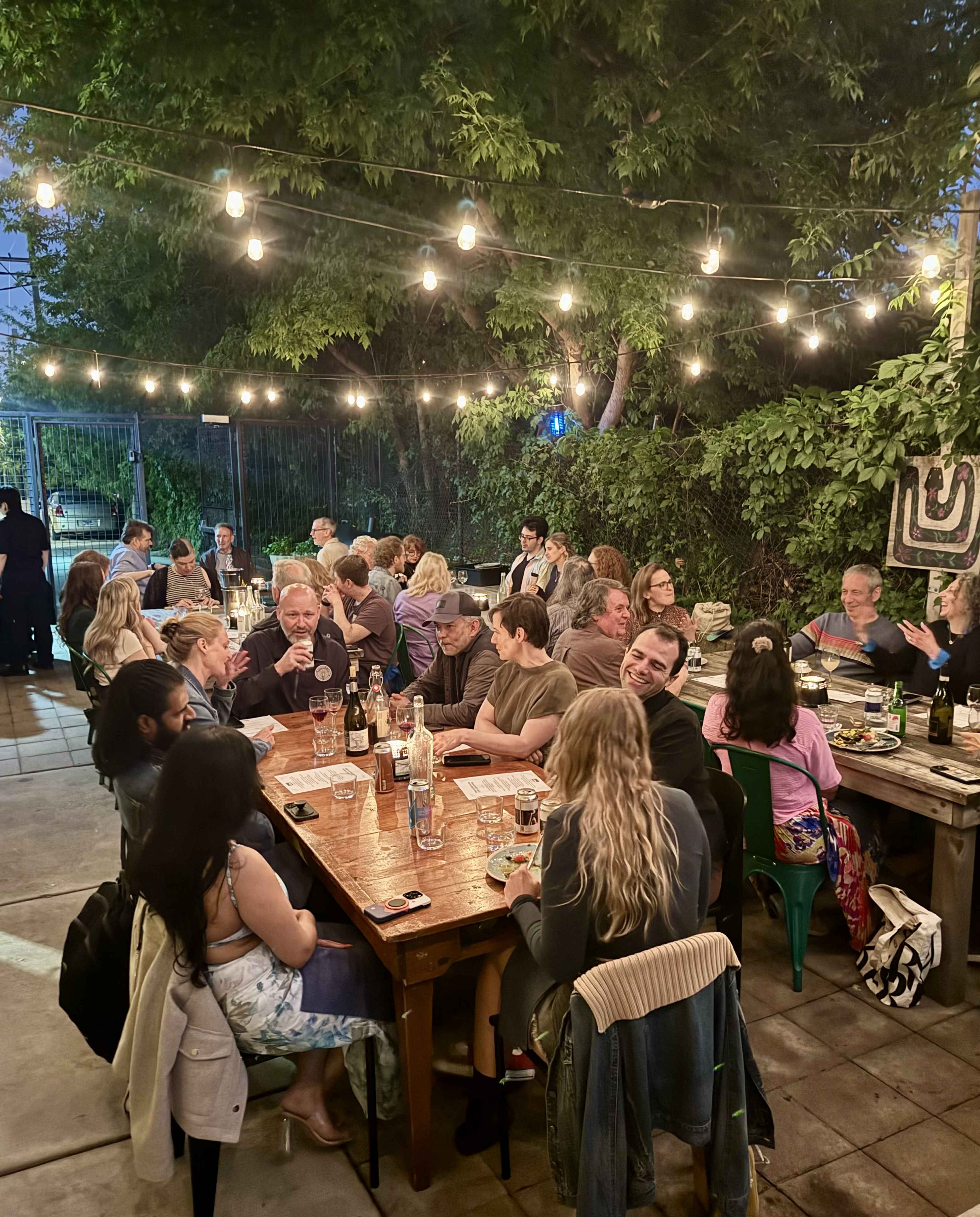 A group of people is seated around several tables outdoors, under string lights, enjoying dinner in a green, leafy environment.