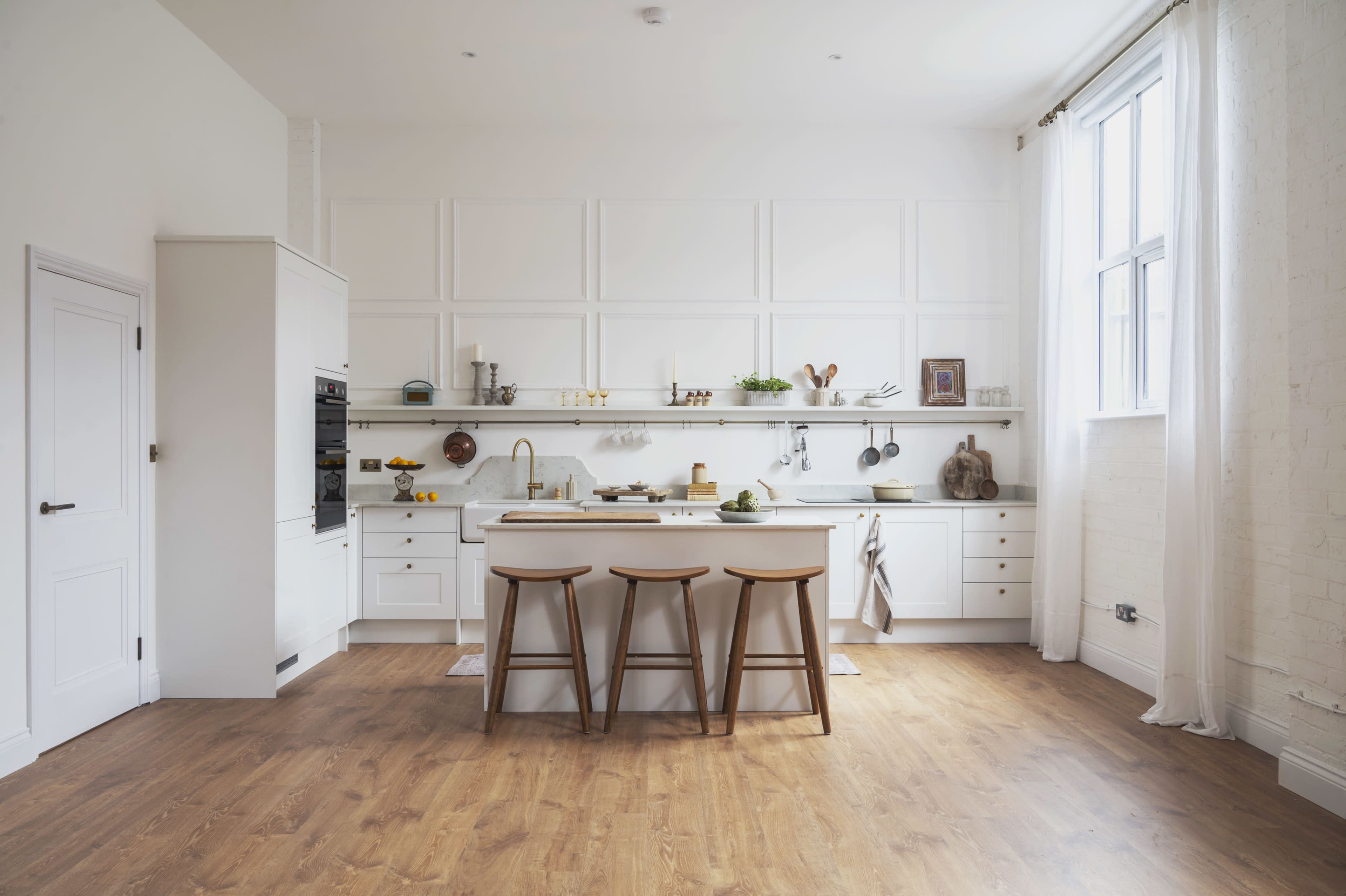 The image shows a modern kitchen with white cabinetry, a central island with three wooden stools, and an array of cooking utensils and decor along the countertop.