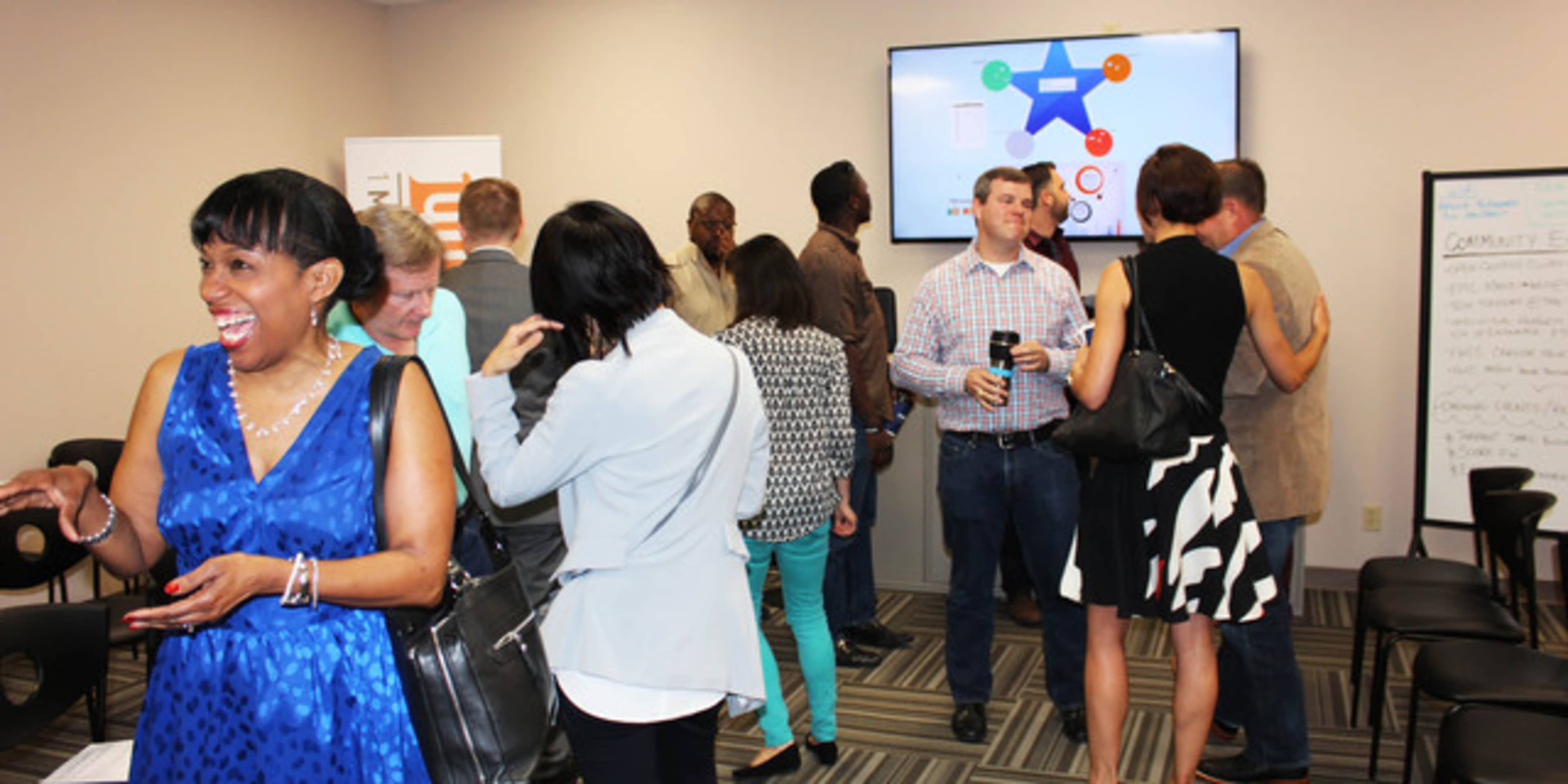 A group of people is interacting in a conference room, with some engaged in conversation while others focus on a presentation displayed on a screen.