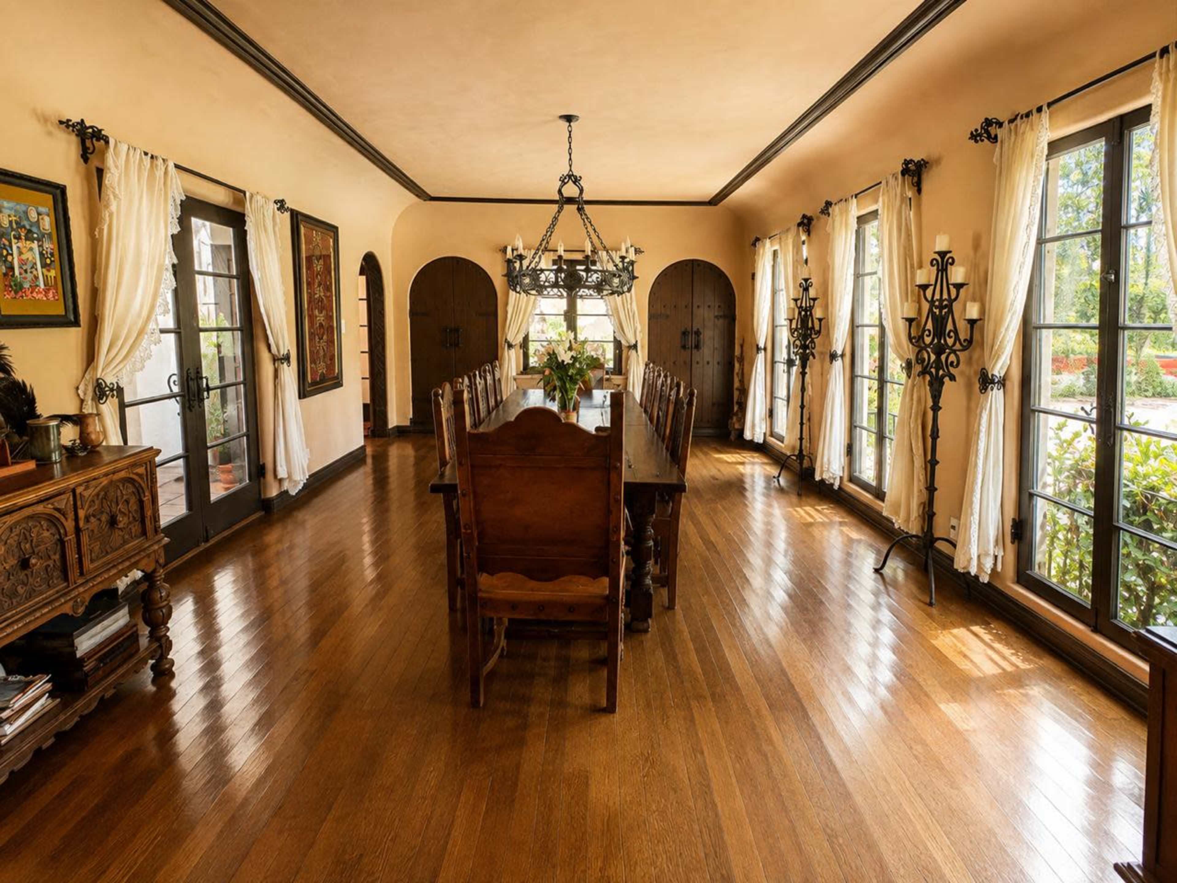 The image shows a spacious dining room featuring a long wooden table surrounded by chairs, large windows with sheer curtains, and decorative elements like a chandelier and wall art.