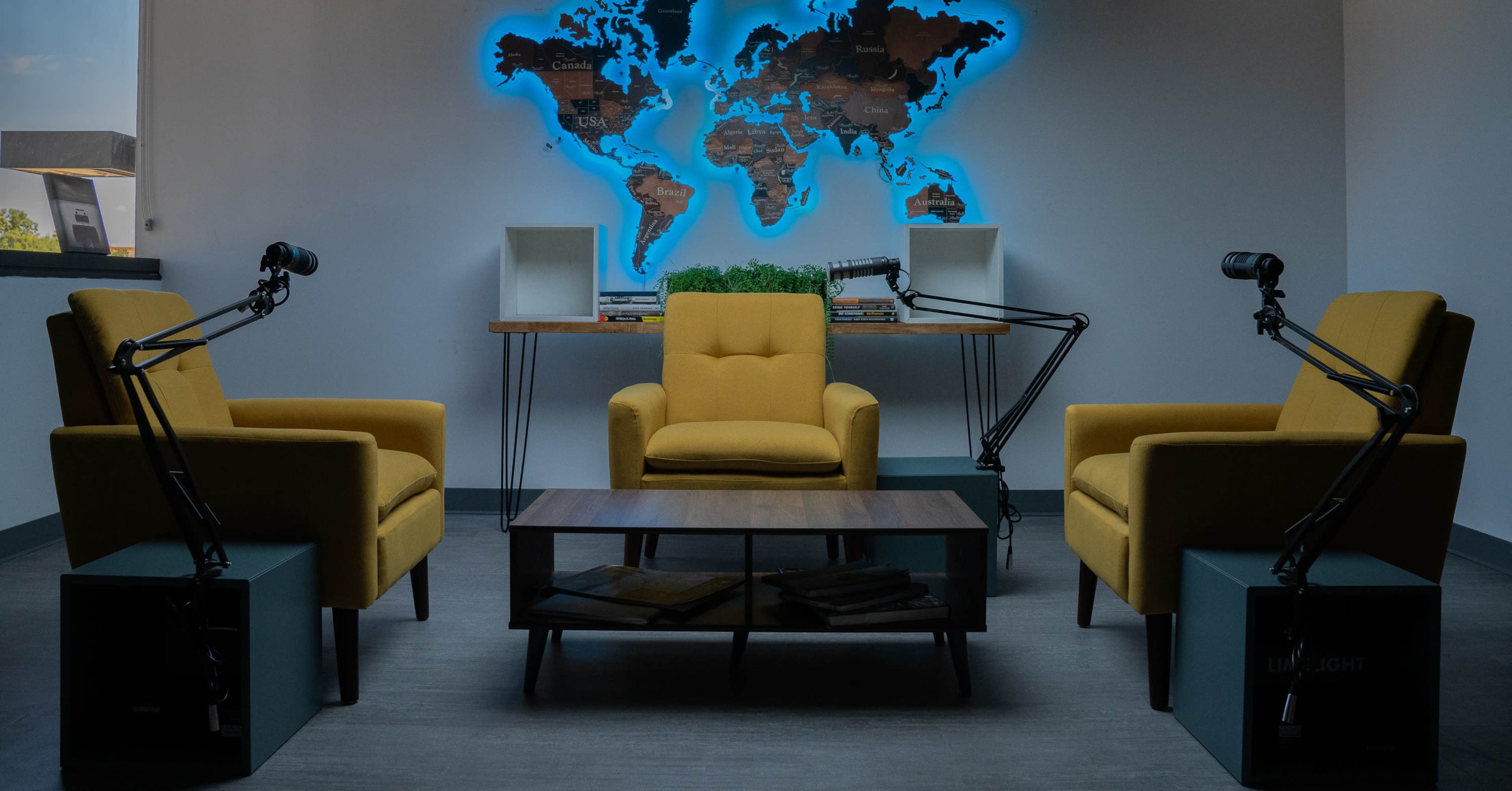A modern podcast studio features three yellow chairs, a wooden coffee table, and a world map illuminated in blue on the wall.