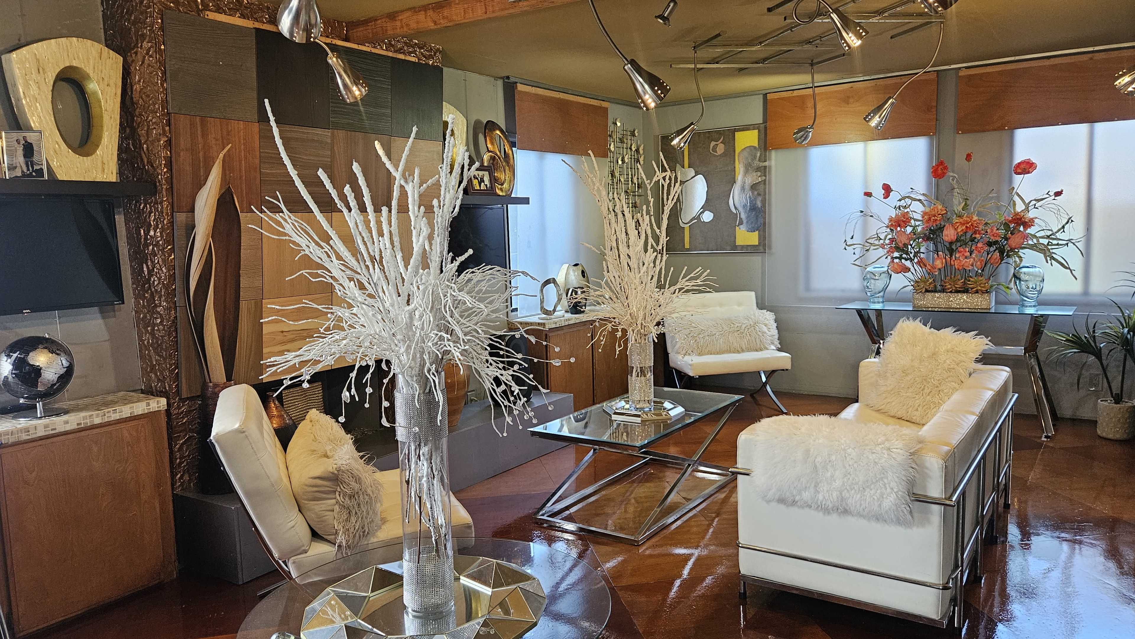 The image features a modern living room decorated with minimalist furniture, including white sofas, metallic accents, and large decorative arrangements of flowers.