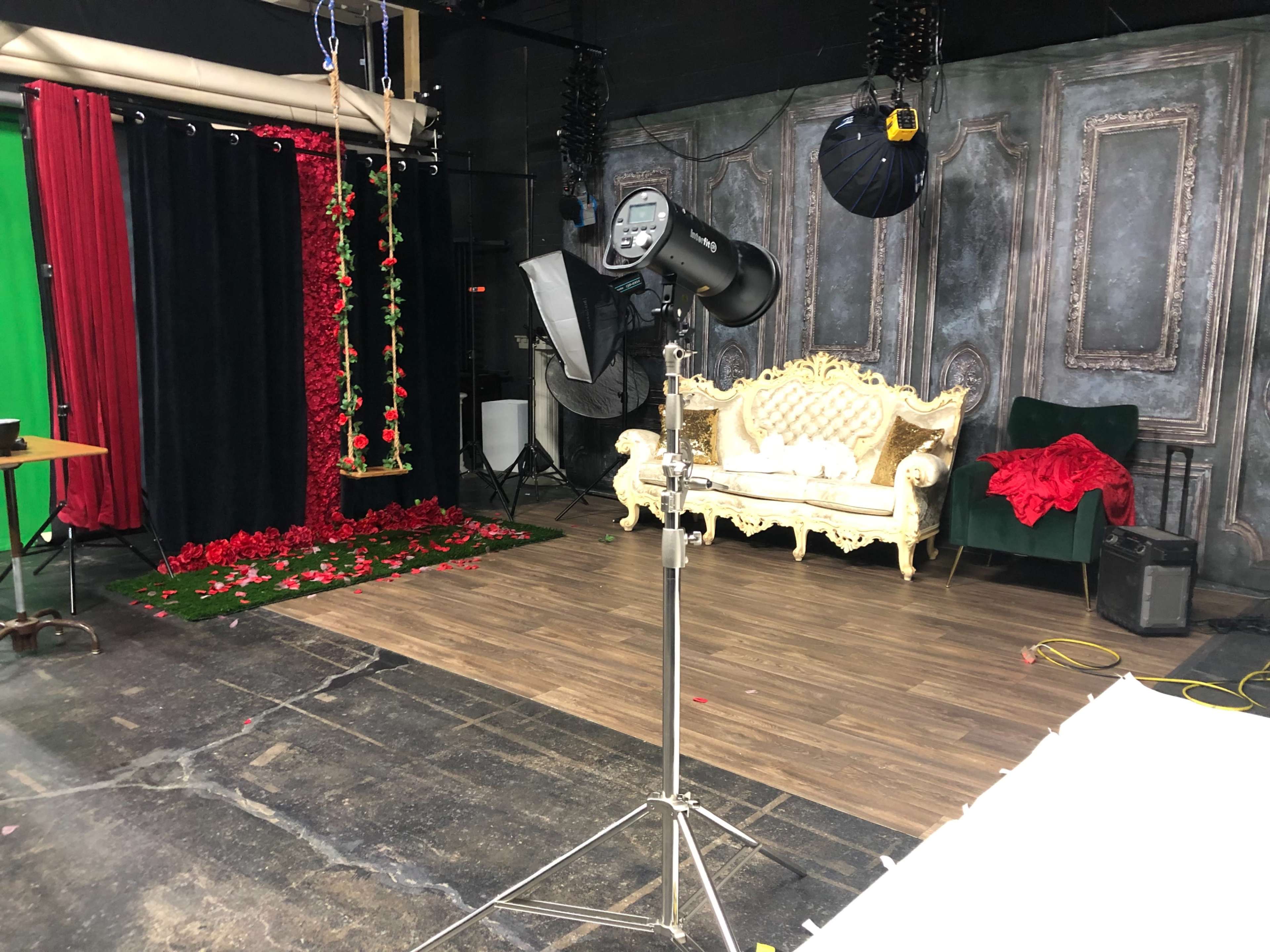 A studio setup with a decorative couch, a swing adorned with roses, and various lighting equipment against a backdrop of draped fabric and a wooden floor.