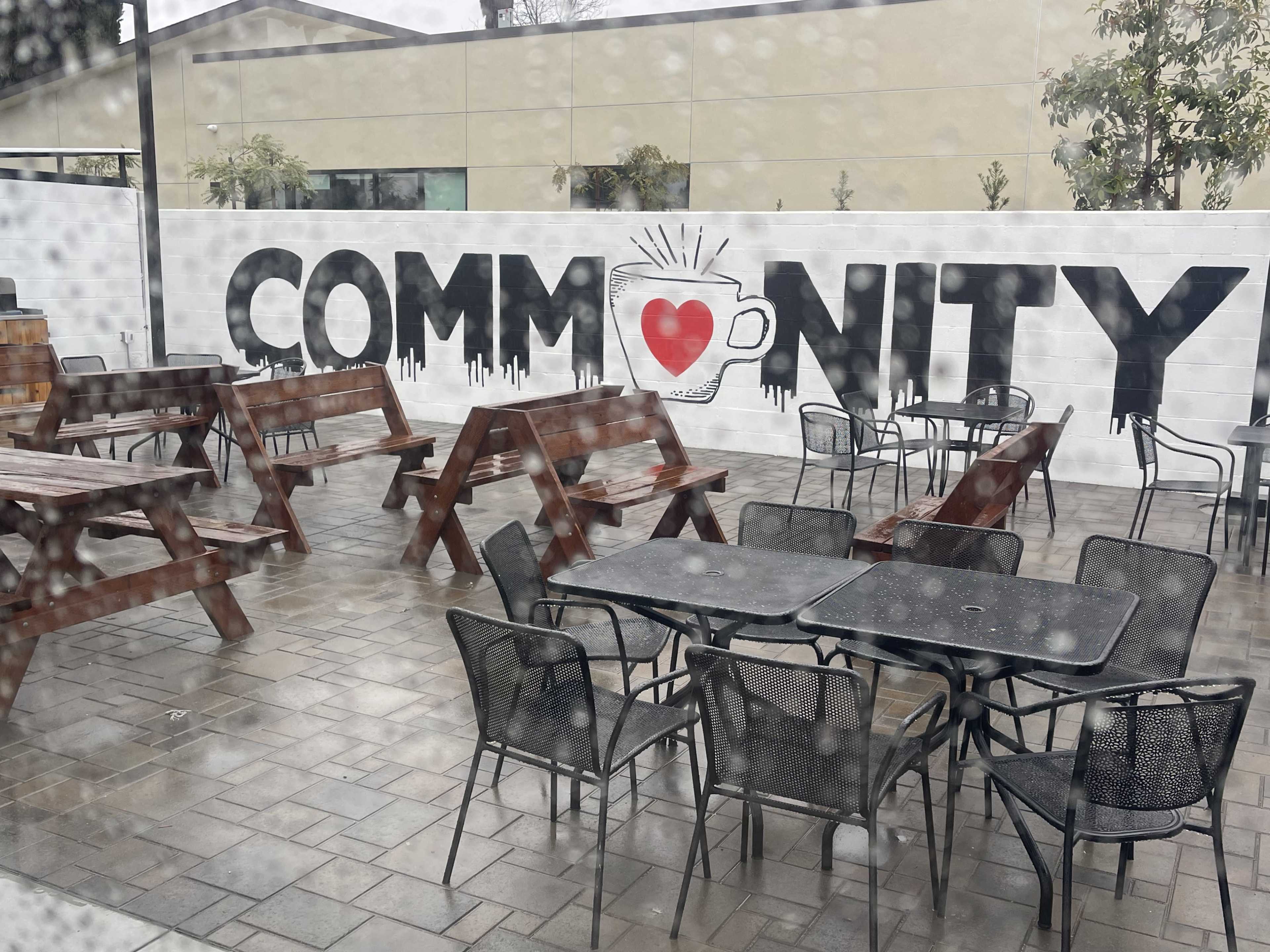 The image shows an outdoor seating area with various tables and benches, featuring a mural that reads "COMMUNITY" with a heart graphic.