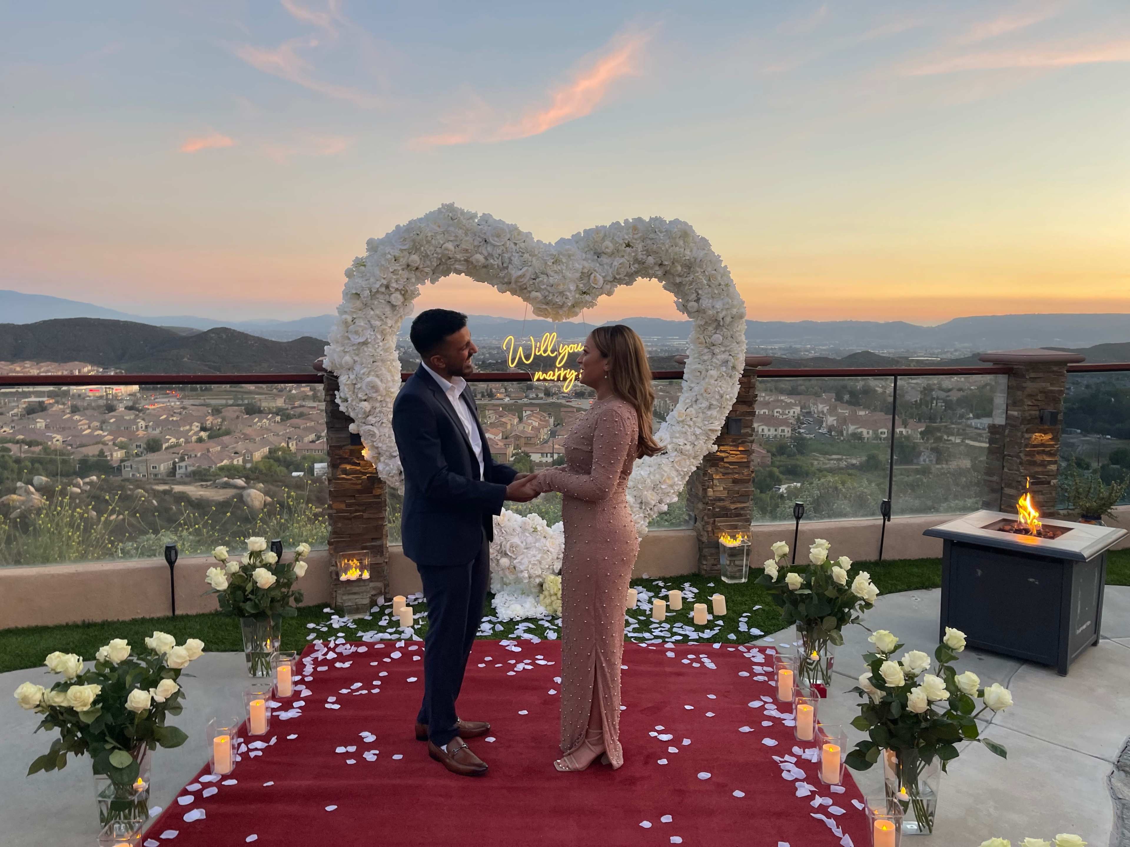A couple stands in front of a heart-shaped floral display at sunset, surrounded by candles and rose petals on a red carpet.
