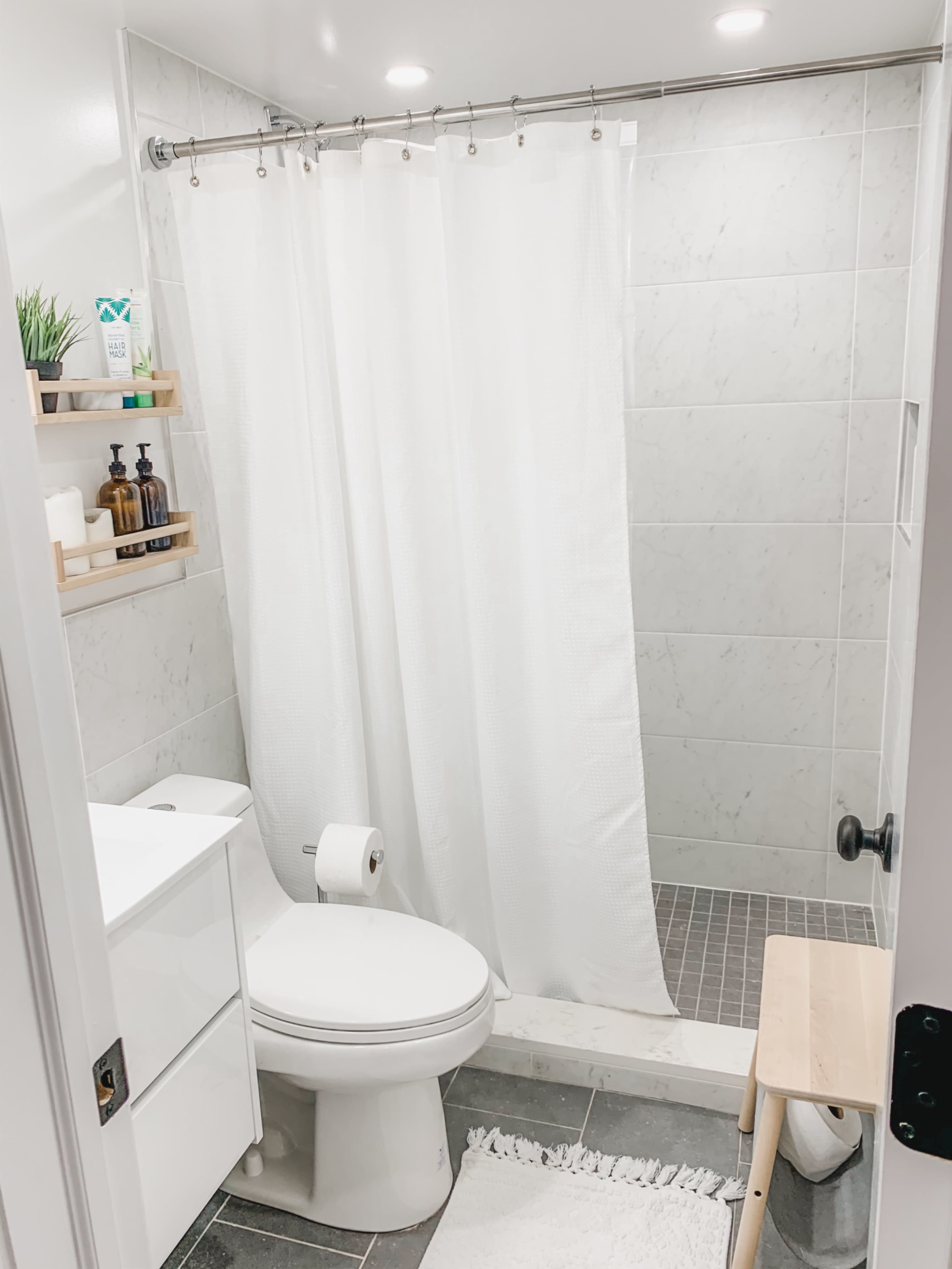 A small bathroom features a toilet, a shower with a glass door, and shelving with various toiletries.