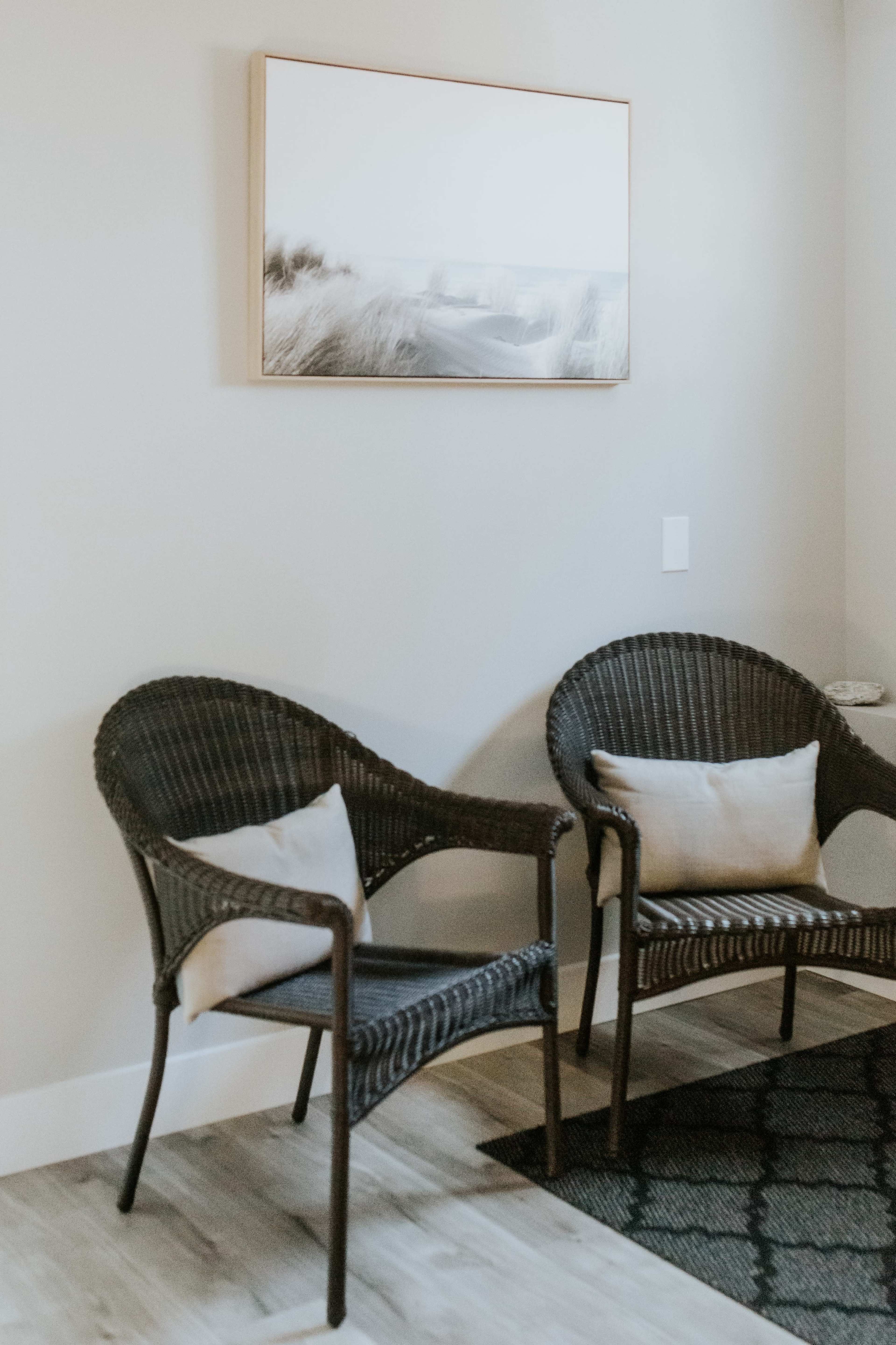 Two wicker chairs with cushions are positioned against a light-colored wall, beneath a framed black-and-white landscape photograph.