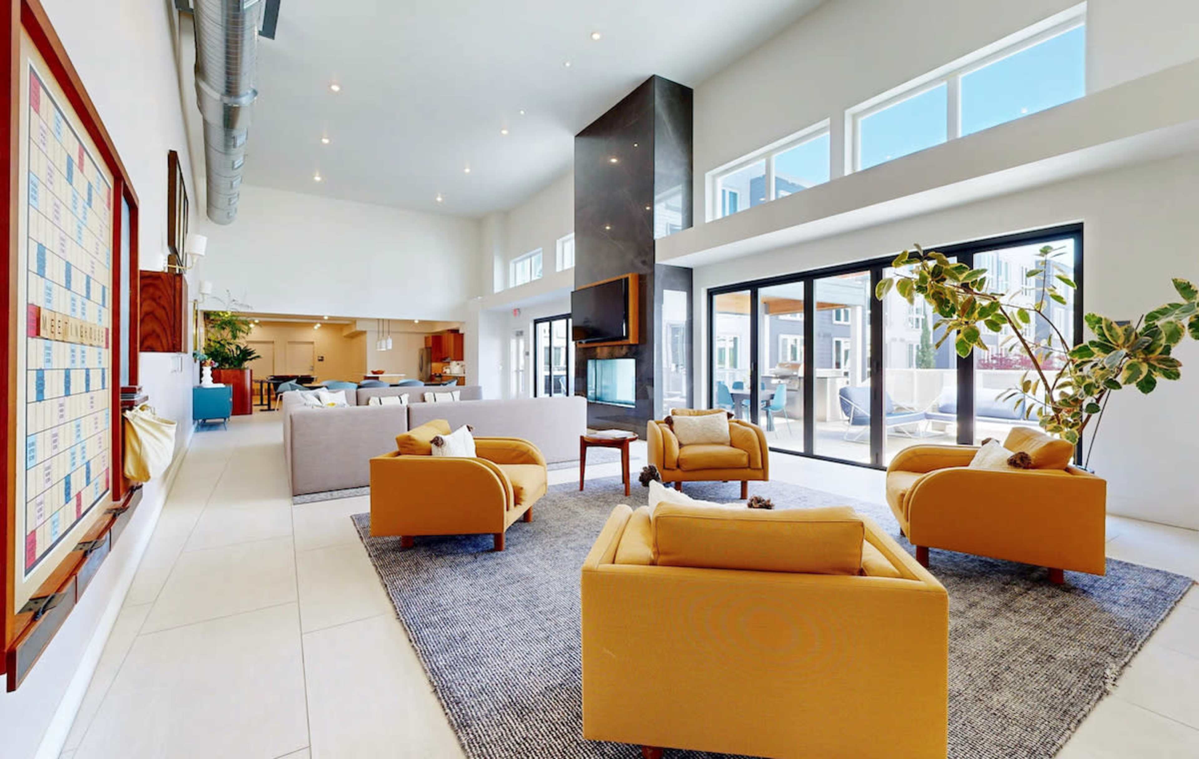 The image shows a spacious modern living area featuring yellow armchairs, large windows allowing natural light, and an open layout with a view of an outdoor space.