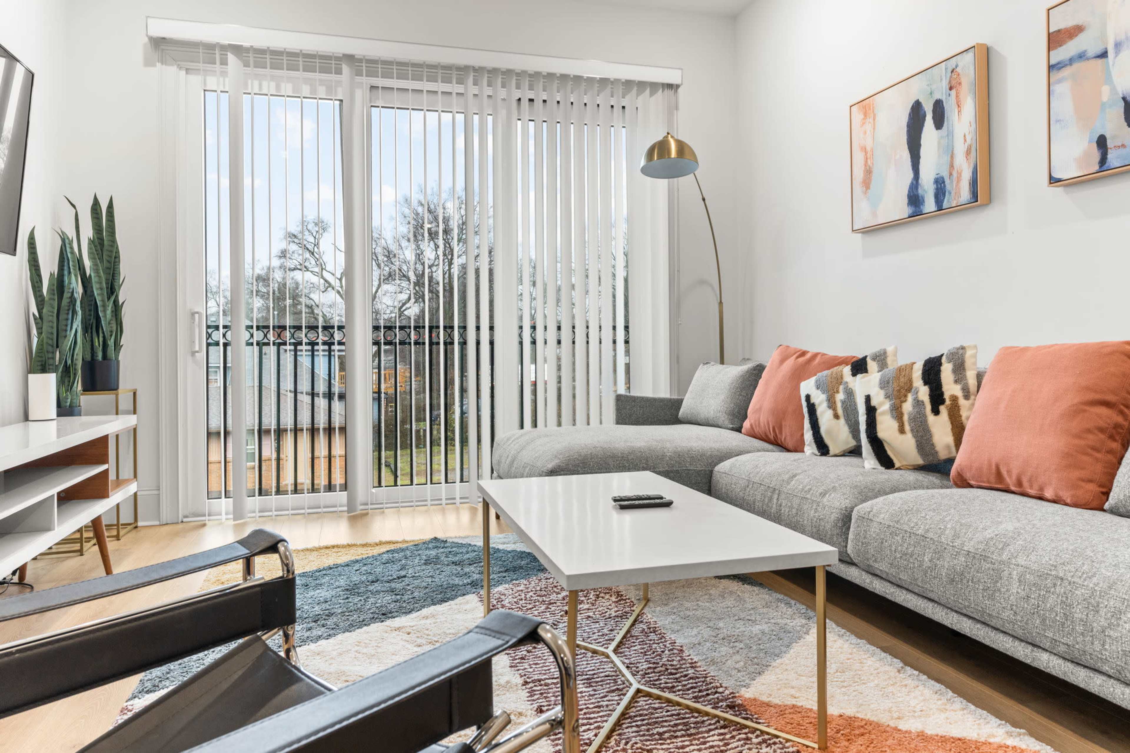 A modern living room features a gray sectional sofa with orange pillows, a white coffee table, a patterned rug, and sliding glass doors leading to a balcony.