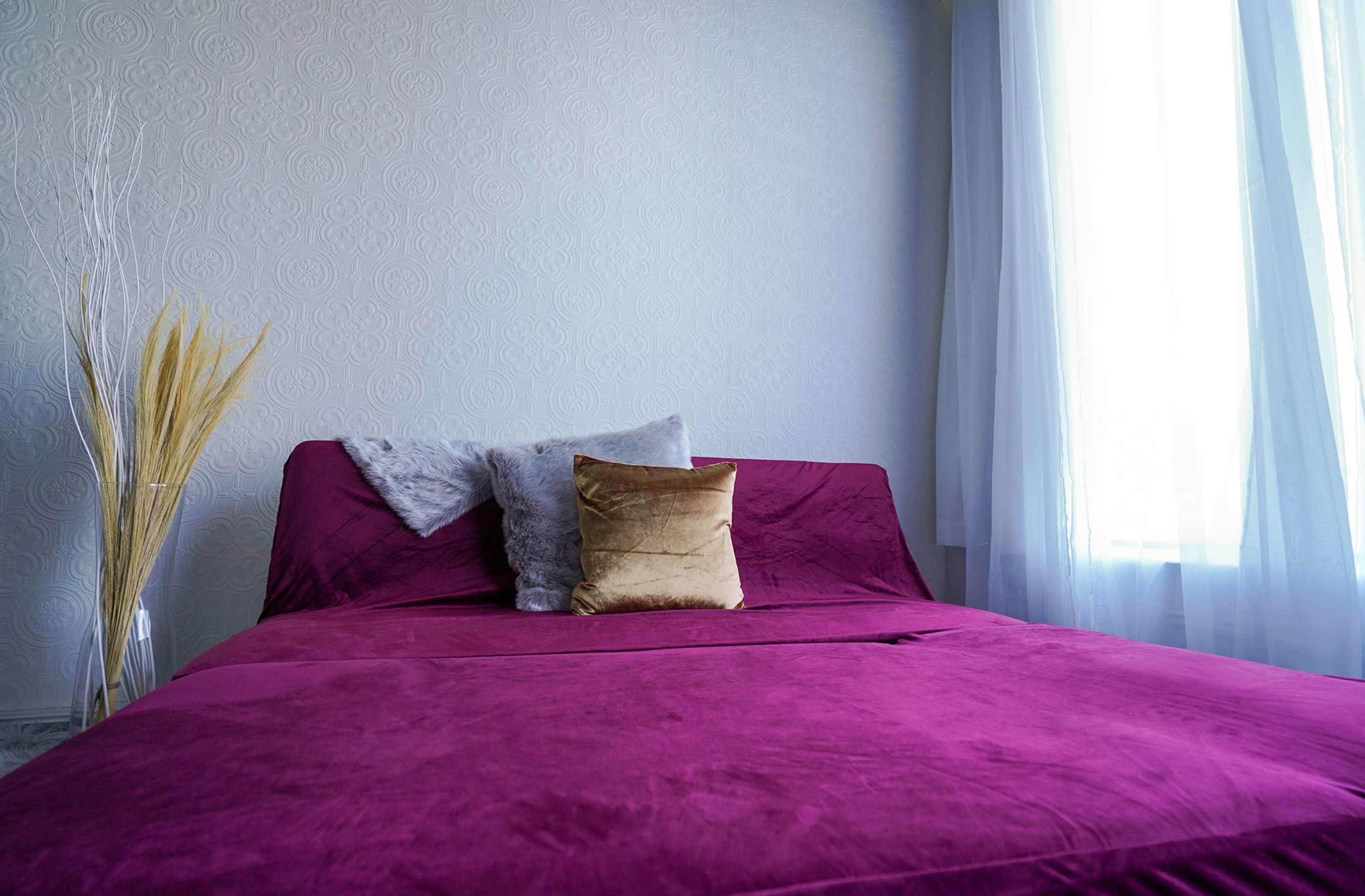 A bed with a deep purple cover and decorative pillows is positioned near a window with sheer white curtains.