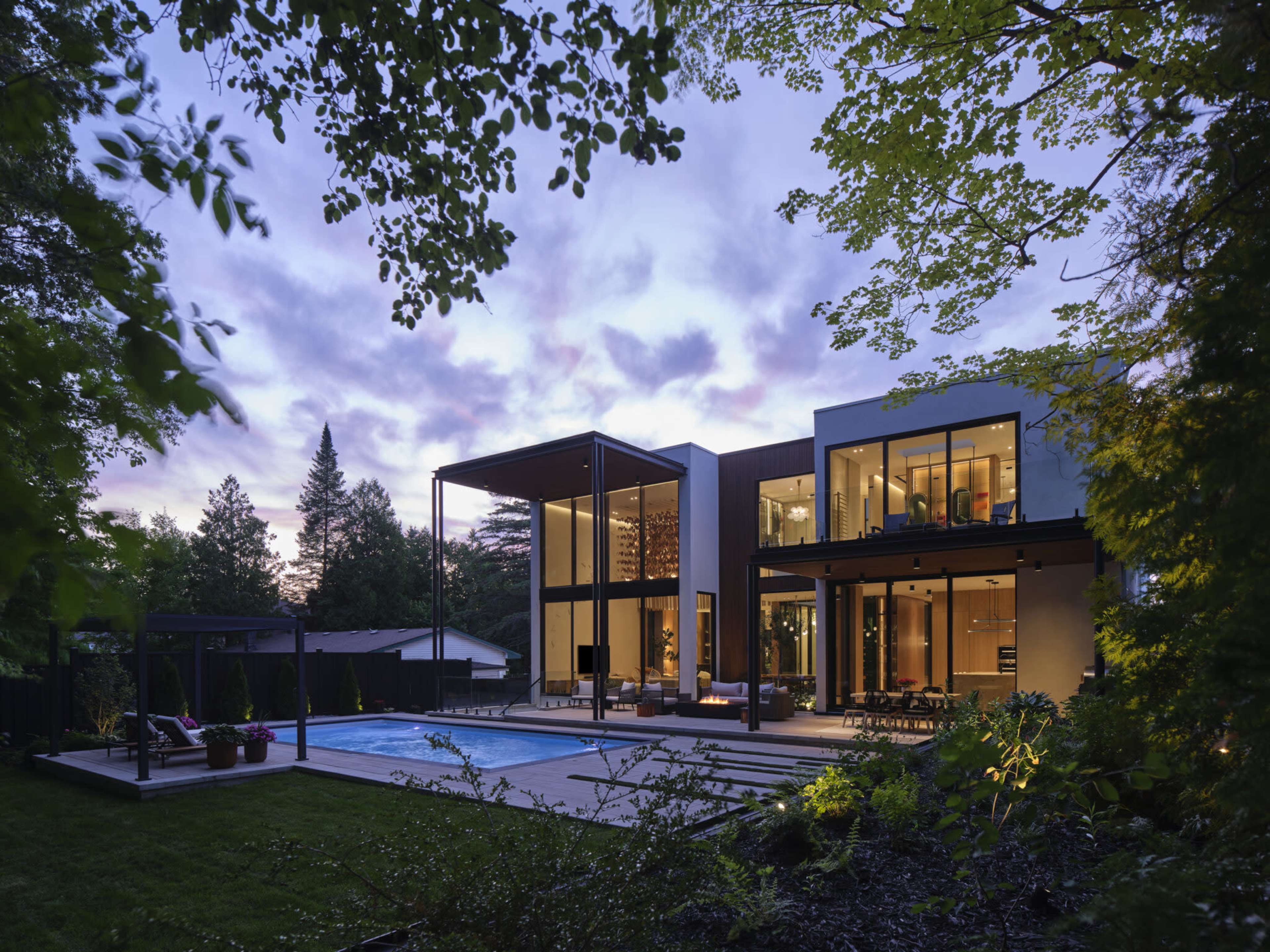 A modern house with large glass windows and a patio overlooks a swimming pool, surrounded by trees and landscaping at twilight.