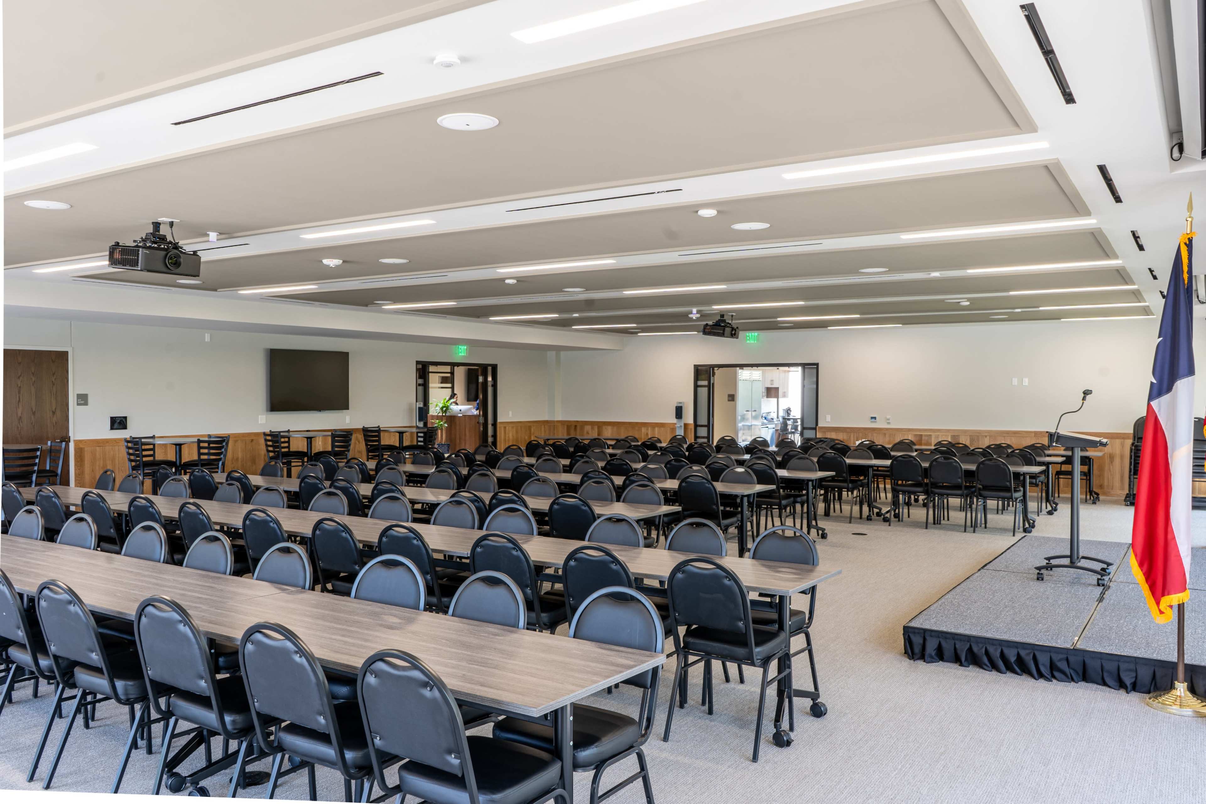 A large meeting room is arranged with multiple rows of black chairs and long tables, featuring a stage area in the front and a Texas flag in the corner.