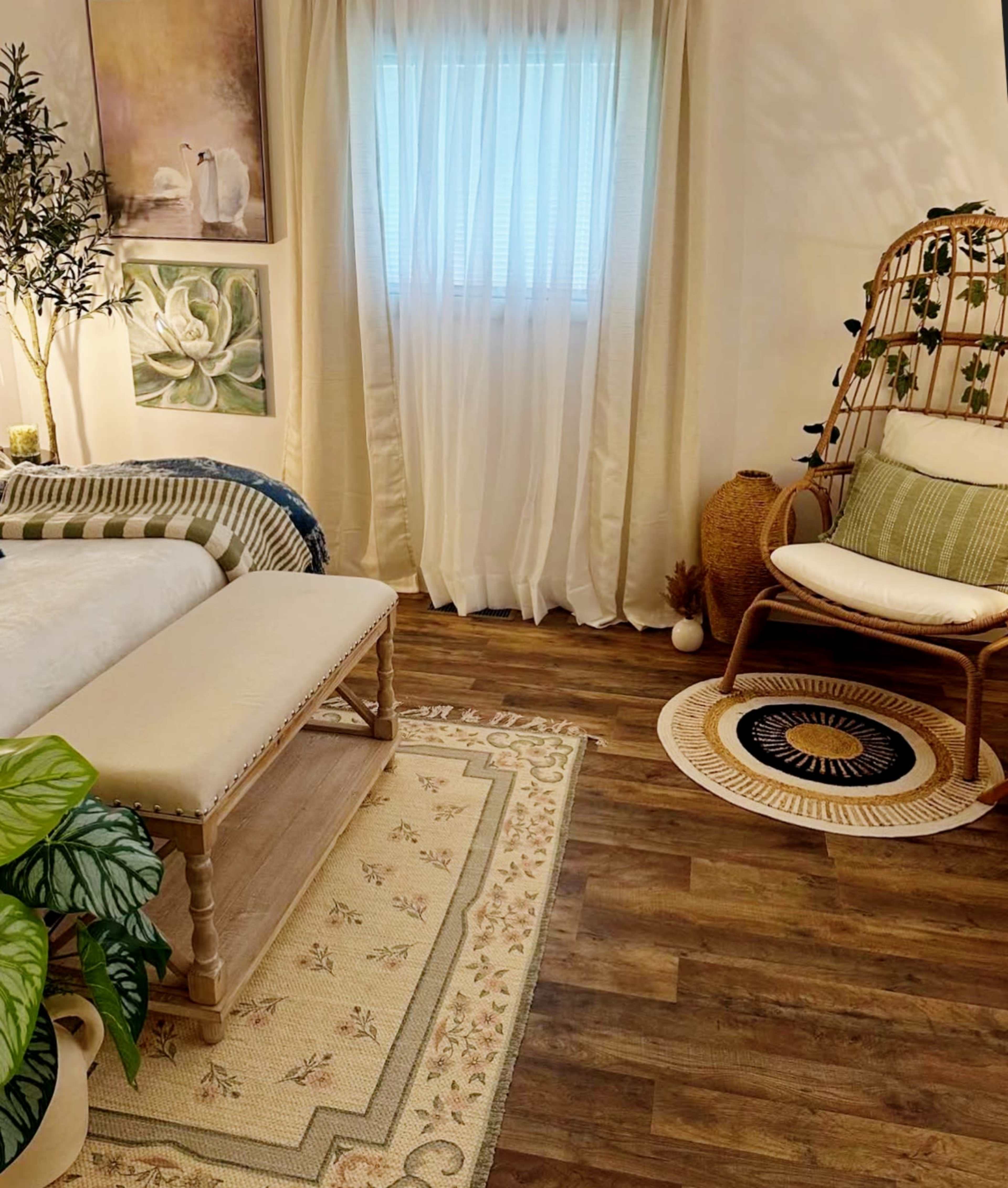 The image shows a cozy living space featuring a bench, a plant, a patterned rug, and a comfortable chair beside a large window with light curtains.