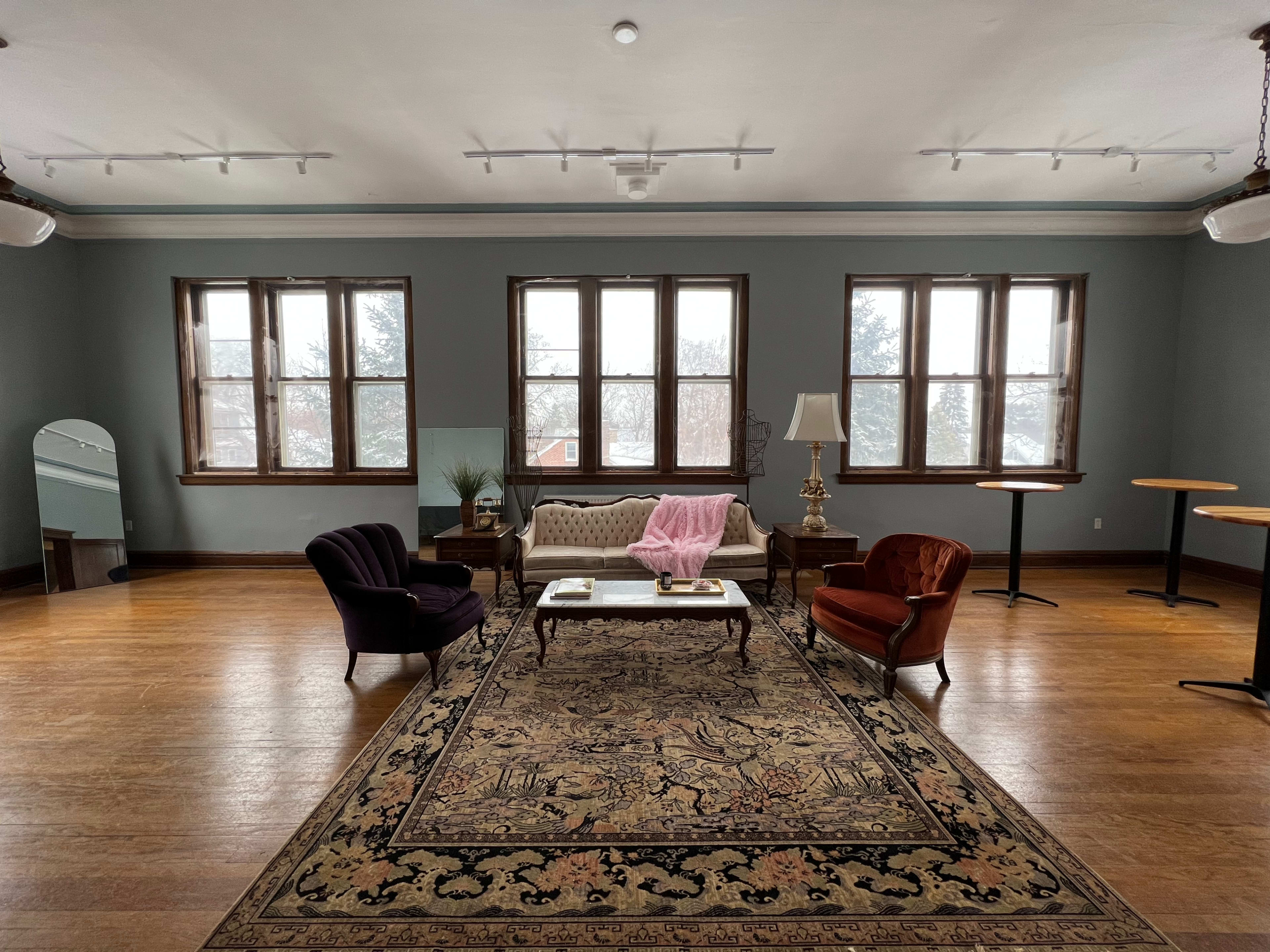 A spacious room features a large area rug with furniture including a couch, two armchairs, a coffee table, and large windows letting in natural light.